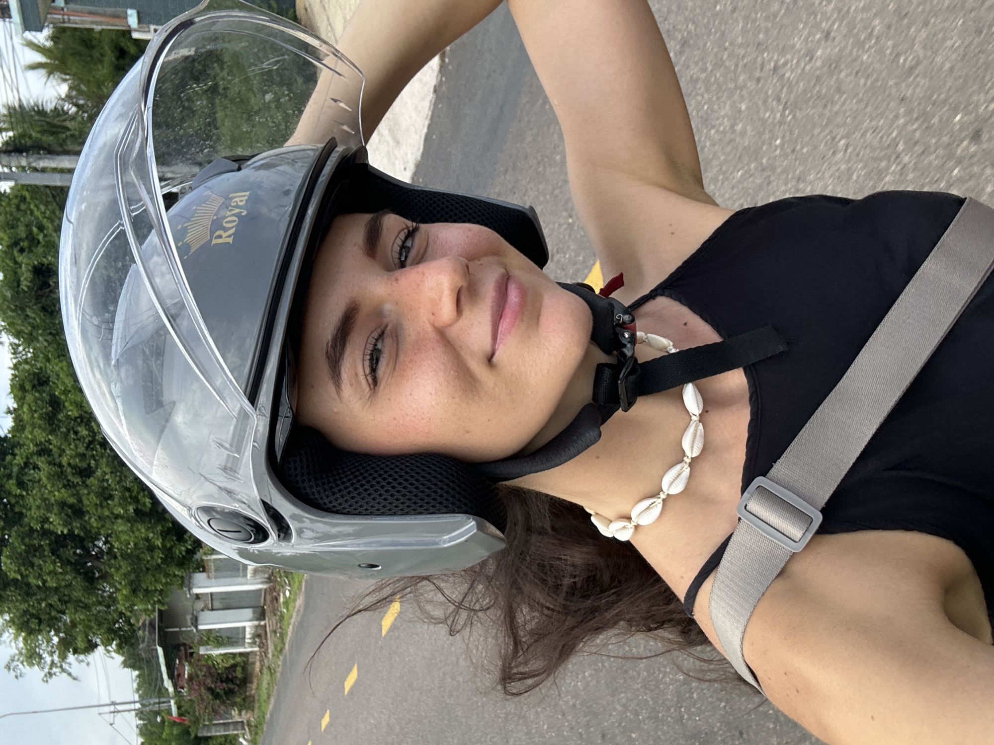 A young woman smiles softly at the viewer in this portrait, captured outdoors in bright daylight. She is depicted from the chest up, wearing a grey open-face motorcycle helmet with a raised clear visor. The helmet features a gold emblem with the word "Royal" and a crown design. Her dark hair is visible below the helmet, framing her face which shows light-colored eyes and freckles. She wears a black top and a distinctive white seashell necklace, along with a grey strap, likely from a bag, across her shoulder.
The background suggests an outdoor setting on a paved road, identifiable by yellow markings on the asphalt. Lush green trees and glimpses of distant structures indicate a tropical or semi-urban environment, consistent with the location of Mũi Né, Vietnam. The sky appears clear, suggesting sunny weather. The image itself is rotated 90 degrees clockwise. The woman's relaxed expression conveys a sense of contentment or a casual moment during her travels, likely on a scooter or motorcycle.