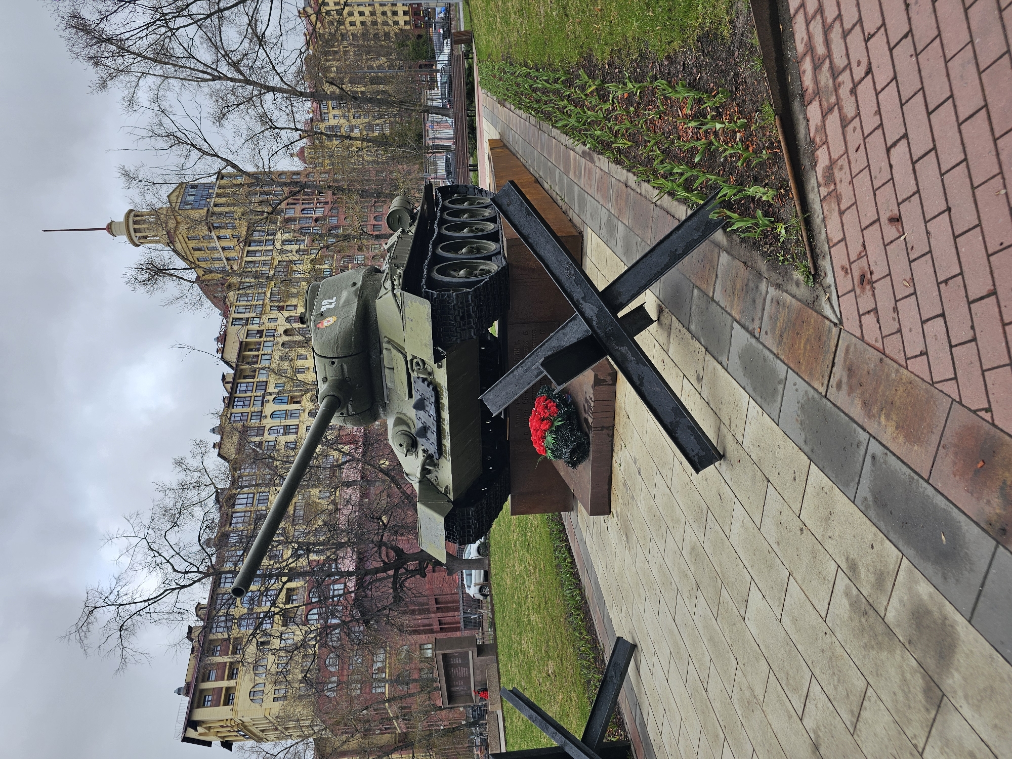 The image depicts a T-34 tank monument located in an urban environment. The tank is positioned on a pedestal behind a wreath and in front of a barrier comprised of black steel anti-tank obstacles. In the background, a multi-story building with a distinctive architectural style and spire rises above leafless trees. The scene is viewed from ground level, with a paved walkway in the foreground bordered by grass and flowerbeds. The sky is overcast, indicating a cloudy day.