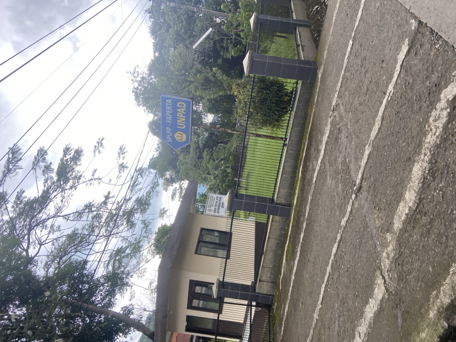 The image shows a paved road in front of a small, beige building with a black metal gate and fence. The gate is closed. Behind the fence is a small, grassy yard. There is a blue sign above the gate, pointing right, which reads "SPLPP - FAPERTA UNPAD". There are many trees and power lines overhead. The image was taken in Baleendah, Indonesia at the coordinates -7.022, 107.658. The scene is empty of people. The ground is asphalt with white painted lines.