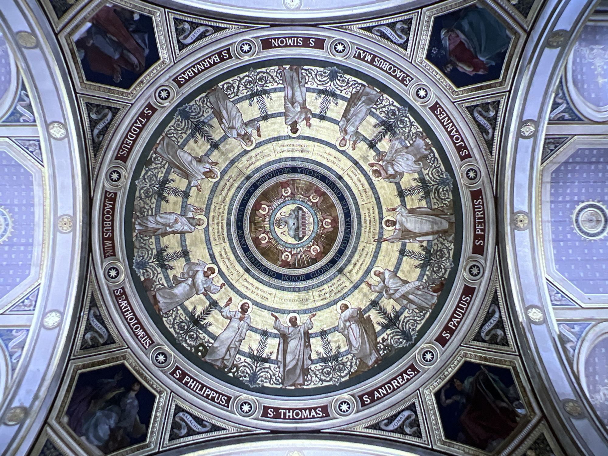 The image shows the ceiling of a church with elaborate fresco paintings. The ceiling is circular and divided into segments, with depictions of figures in robes, some with halos, arranged in a circular pattern. In the center of the ceiling, there is a medallion with a religious scene, possibly depicting the Lamb of God. Around the central medallion, there are concentric circles with inscriptions in Latin. The overall style is ornate and appears to be of Renaissance or Baroque origin. The paintings are rendered in warm, earthy tones with gold accents. This is the ceiling of the Basilica di San Zeno Maggiore in Verona, Italy, specifically the ceiling of the main nave.
