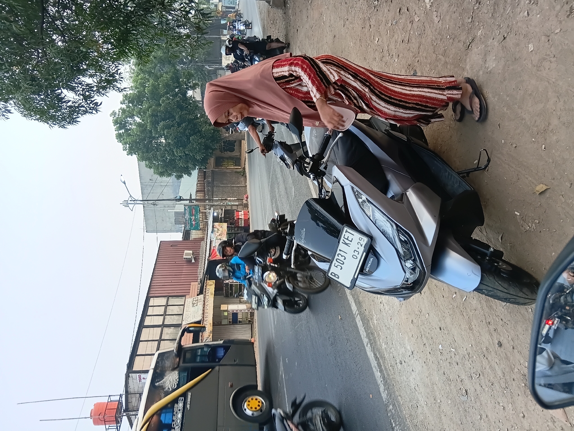A woman wearing a pink hijab and a striped dress stands on a dirt roadside next to a silver Honda motorcycle in Bekasi, Indonesia. The motorcycle is parked and facing towards the viewer. A white plate with the inscription "B 5031 KET 03.29" is visible on the front of the motorcycle. In the background, traffic moves along a paved road, including a bus and several other motorcycles. Buildings, trees, and utility poles line the street. The sky is overcast.