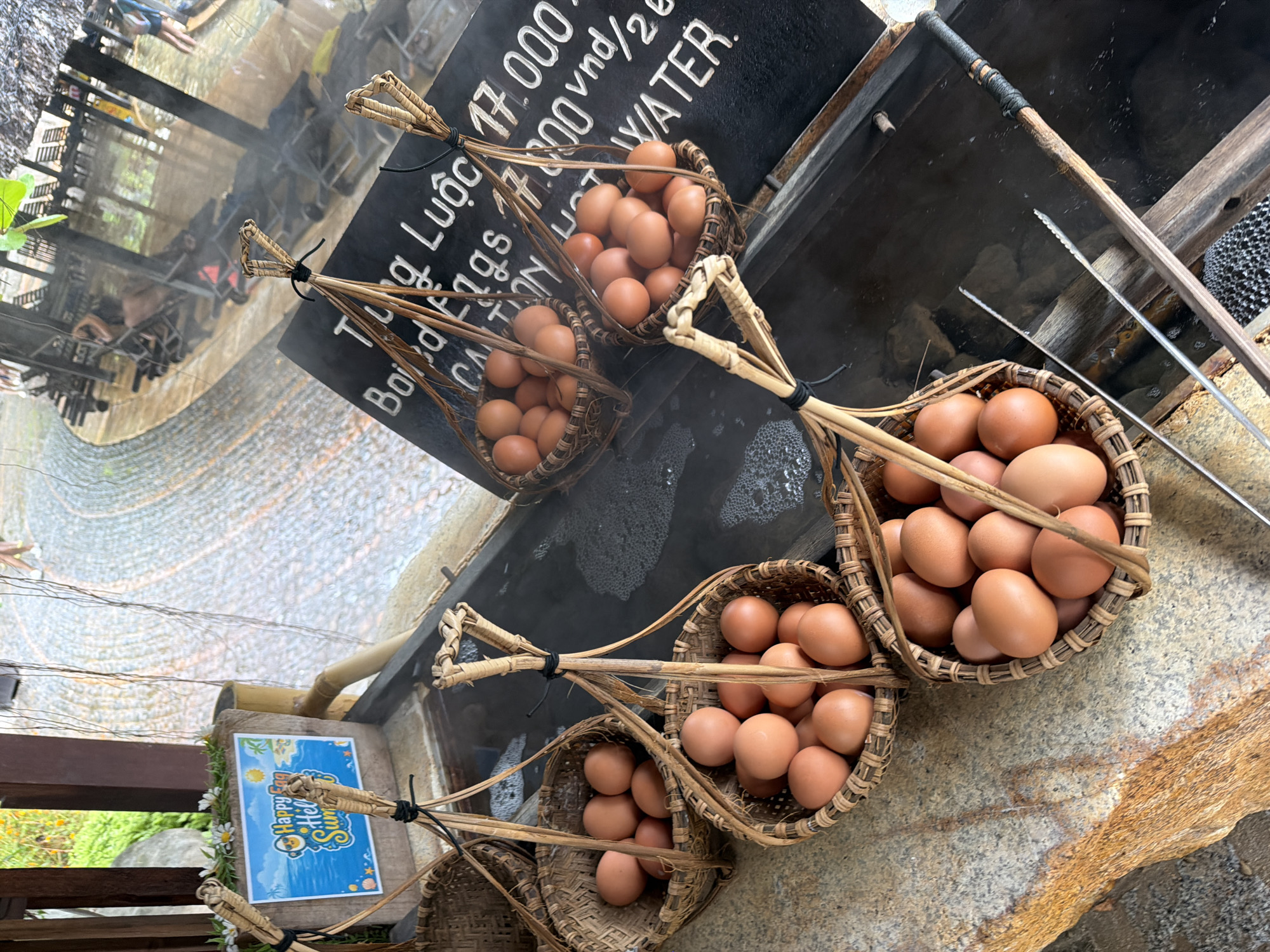 This is a close-up, eye-level shot of baskets of eggs placed near a hot spring in Nha Trang, Vietnam. The eggs are light brown and are contained in woven baskets, which are suspended by bamboo handles. The baskets appear to be ready for boiling in the hot spring, as evidenced by the steam rising from the dark water in the background and the presence of metal tongs. A sign with Vietnamese text and prices is visible behind one of the baskets. The overall atmosphere suggests a tourist attraction where visitors can boil their own eggs in the natural hot springs.