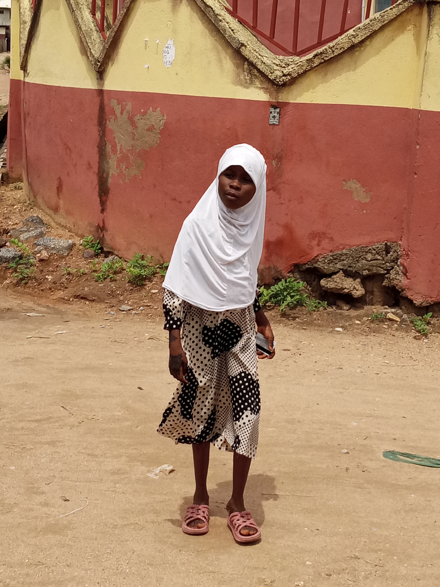 A young girl stands on a dirt road in front of a two-tone building in Unknown, Unknown. The girl is wearing a white headscarf and a black and white polka dot dress. She is also wearing pink sandals. The building behind her is painted yellow on top and red on the bottom. There is some peeling paint on the red section of the building. There is also some vegetation growing along the base of the building. The dirt road is dry and dusty.