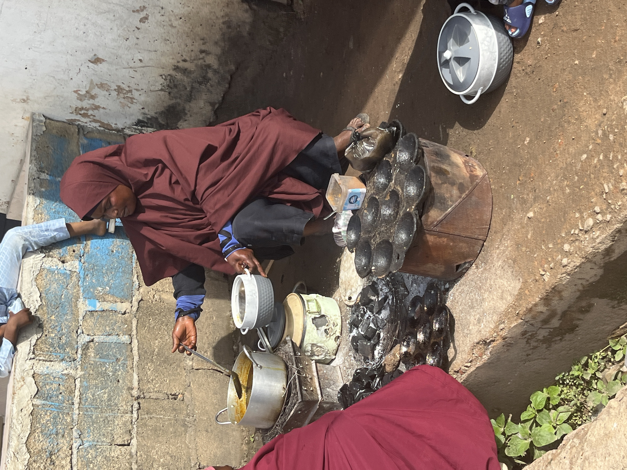In Jos, Nigeria, a woman wearing a maroon hijab and dark pants is crouched down, cooking over an open fire. She is using a metal ladle to stir food in a large pot, which is placed on a makeshift stove. Next to this stove are several smaller cooking vessels. To her right, a grey pot with a lid sits on the ground. The ground itself is composed of dirt and small stones. A child in light blue patterned clothing is visible in the background, sitting on a concrete ledge. The wall behind the woman is made of concrete blocks painted in blue and white. There is also a partially visible figure on the left wearing a maroon garment.