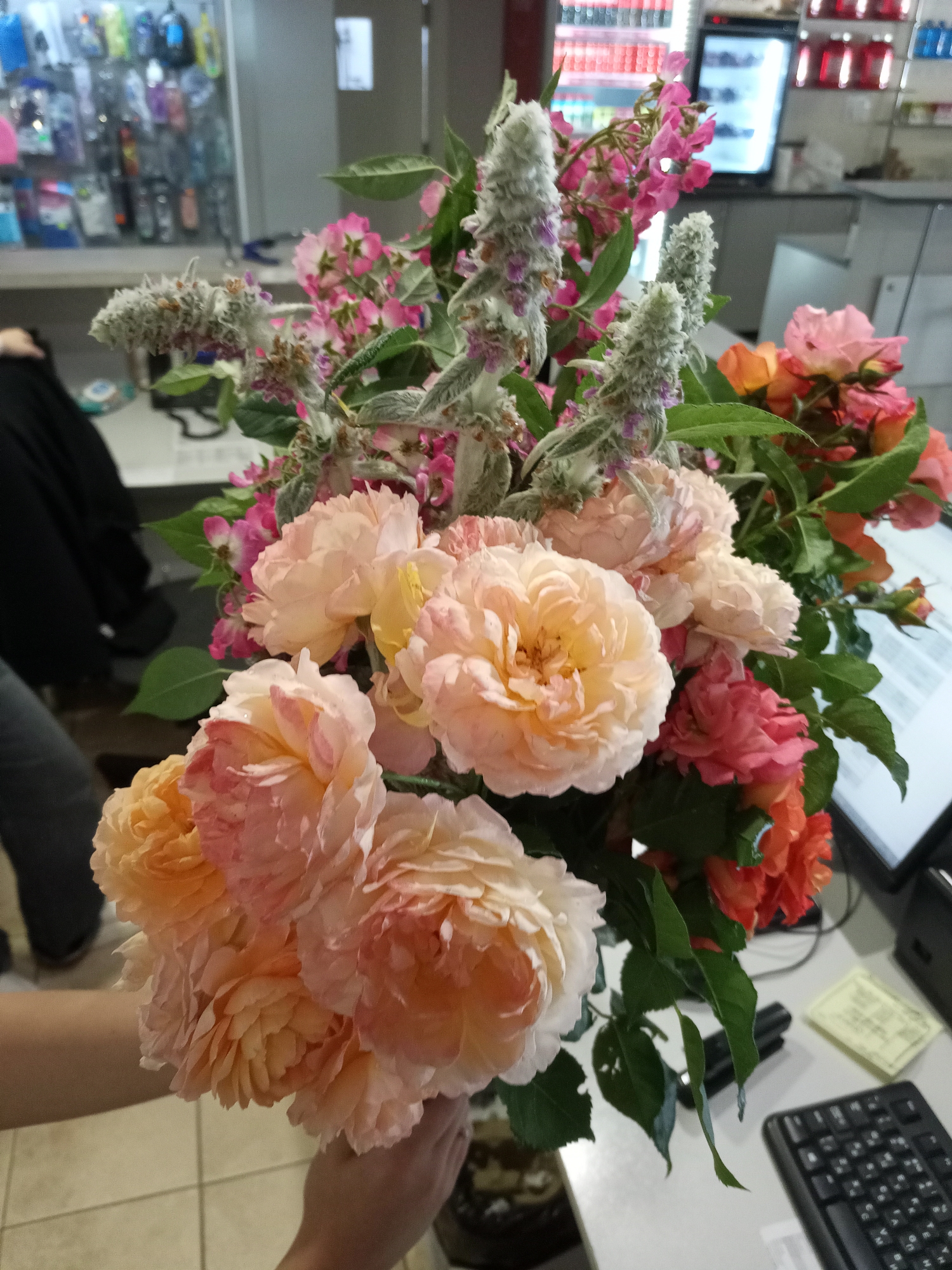 A bouquet of flowers, primarily roses in shades of peach, pink, and orange, is held in the foreground by a person's hands. The roses are large and fully bloomed, with many delicate petals. Interspersed with the roses are spiky, fuzzy plants with small purple flowers and delicate pink flowers. The background is a bit blurry, but appears to be an indoor setting, possibly an office or store. There are shelves stocked with various items, including what looks like bottles and packaged goods. A computer keyboard and a yellow sticky note are visible on a desk in the background. The lighting suggests it could be daytime, but it's difficult to determine the exact time. The overall impression is one of a colorful and vibrant display of flowers.