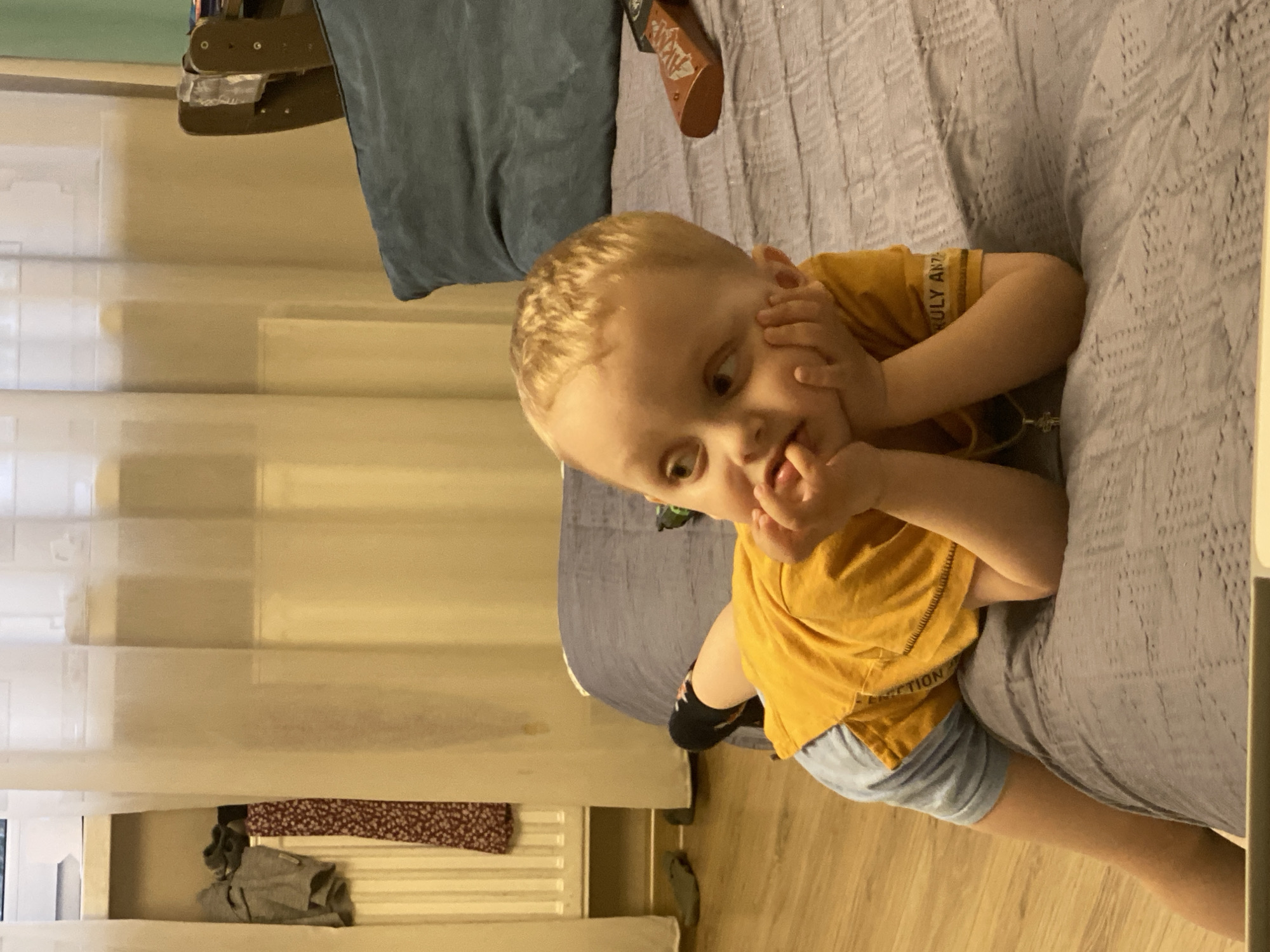 A blond toddler in a mustard-yellow t-shirt and blue shorts is lying on a gray textured bedspread. The child has their hands on their face, fingers near their mouth. Part of a blue pillow is visible behind the child. In the background, a window with closed blinds is to the left, and what appears to be clothing is beneath the window. The floor is wood. There is no indication of the location being Yekaterinburg, Russia.
