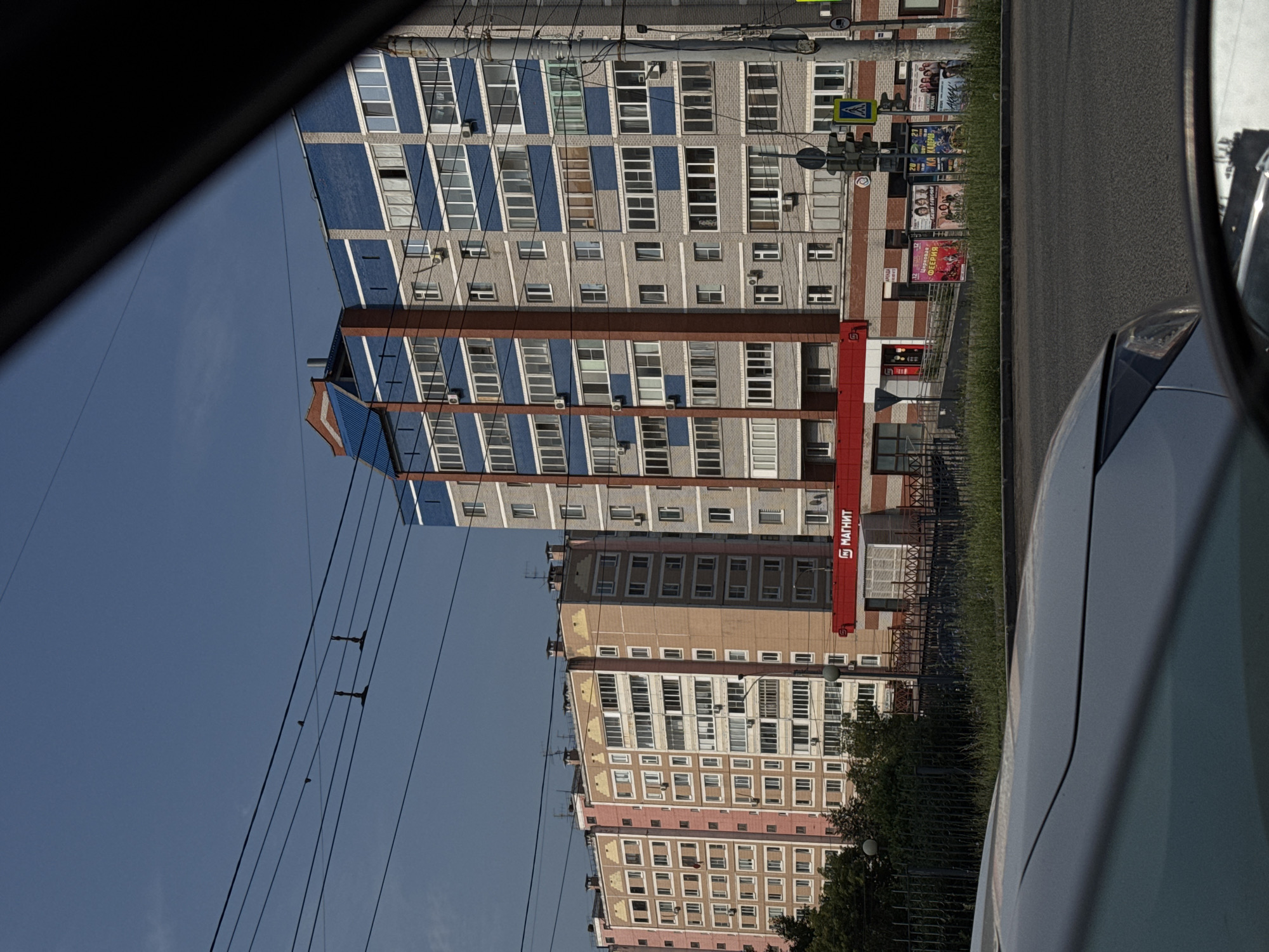 The image shows a view from inside a car looking out at a street scene in Izhevsk, Russia. In the background, there are several tall apartment buildings with blue and pink facades. One of the buildings has a red awning with the word "МАГНИТ" (Magnit), a Russian supermarket chain, visible. There are traffic lights and advertisements on poles along the street. The sky is a clear blue. The foreground of the image is dominated by the hood and side mirror of the car from which the photo is taken.