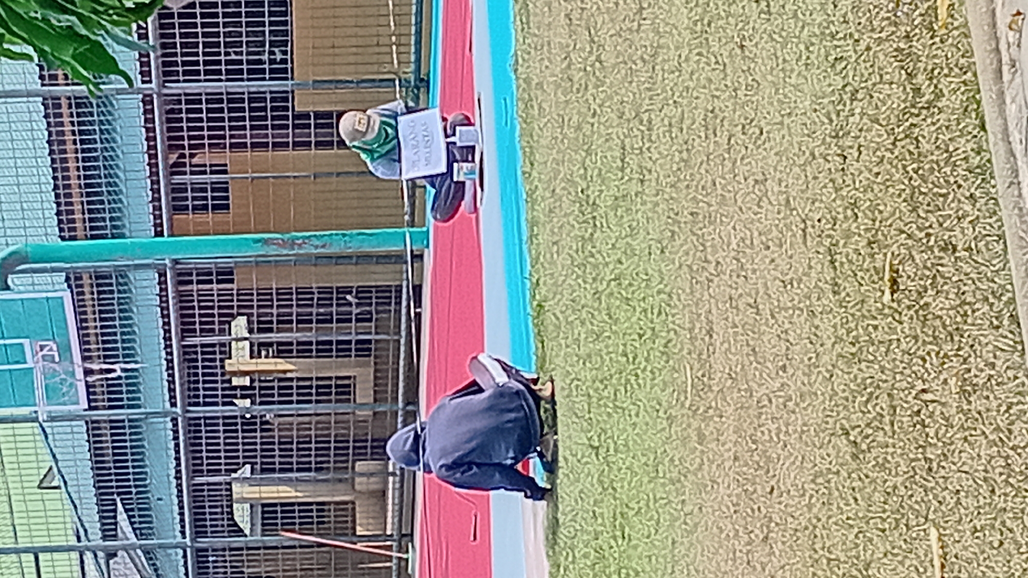 The image depicts two individuals on a red and blue court, likely a basketball court, with a grassy area in the foreground. In the background, a building with a green metal roof and barred windows is visible, suggesting a facility with security measures. A basketball hoop is mounted on a pole.
The person on the left is wearing a dark hooded jacket and is crouched down on the grass, facing away from the camera. A red pole or stick is visible near them.
The person on the right is sitting cross-legged on the court, wearing a light-colored cap and a green scarf or face covering. They are looking down at a white sign with text on it, which appears to be a prohibition sign. Two white cups or containers are placed in front of the sign.
The scene appears to be outdoors in a public area, possibly a school or sports complex, given the basketball court and the surrounding structures. The lighting suggests it is daytime. The image is somewhat pixelated, making fine details challenging to discern.
Without additional context or information specific to the city of Jambi, Indonesia, it is not possible to pinpoint the exact location within the city. However, the presence of a basketball court and a building with a gated facade could indicate a public park, a school, or a recreational facility within Jambi.