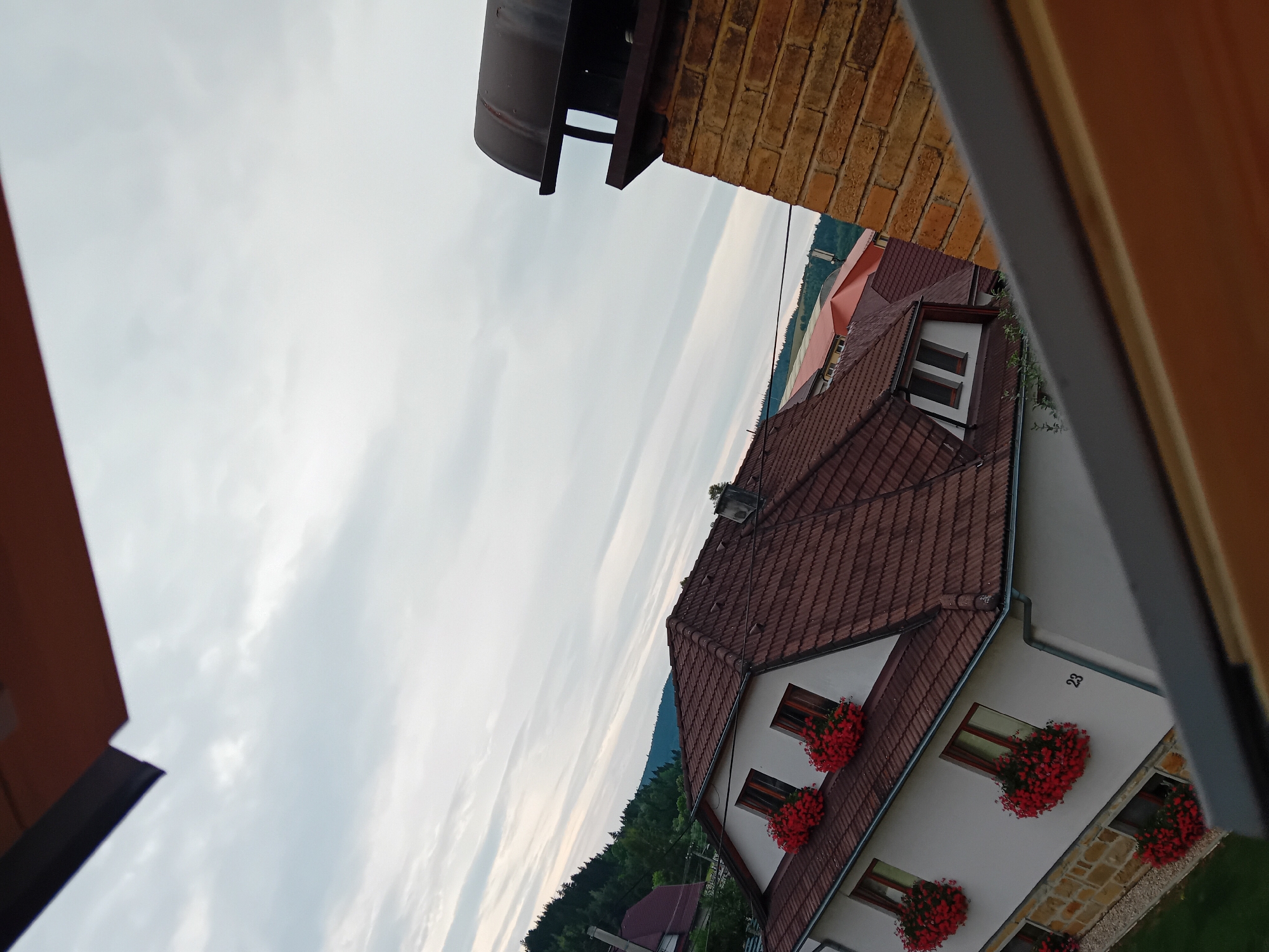 The image captures a view of houses in Zuberec, Slovakia, under an overcast sky. The foreground features elements of a wooden structure, likely part of a balcony or roof overhang, framing the scene. In the mid-ground, a white house with a red-tiled roof is prominent, adorned with numerous window boxes filled with vibrant red flowers. The house is numbered "23". Adjacent to this house, a chimney constructed of orange bricks stands tall. Beyond these structures, a dense forest covers rolling hills, and other buildings with red roofs are visible in the distance. The overall impression is a residential area nestled within a natural landscape.