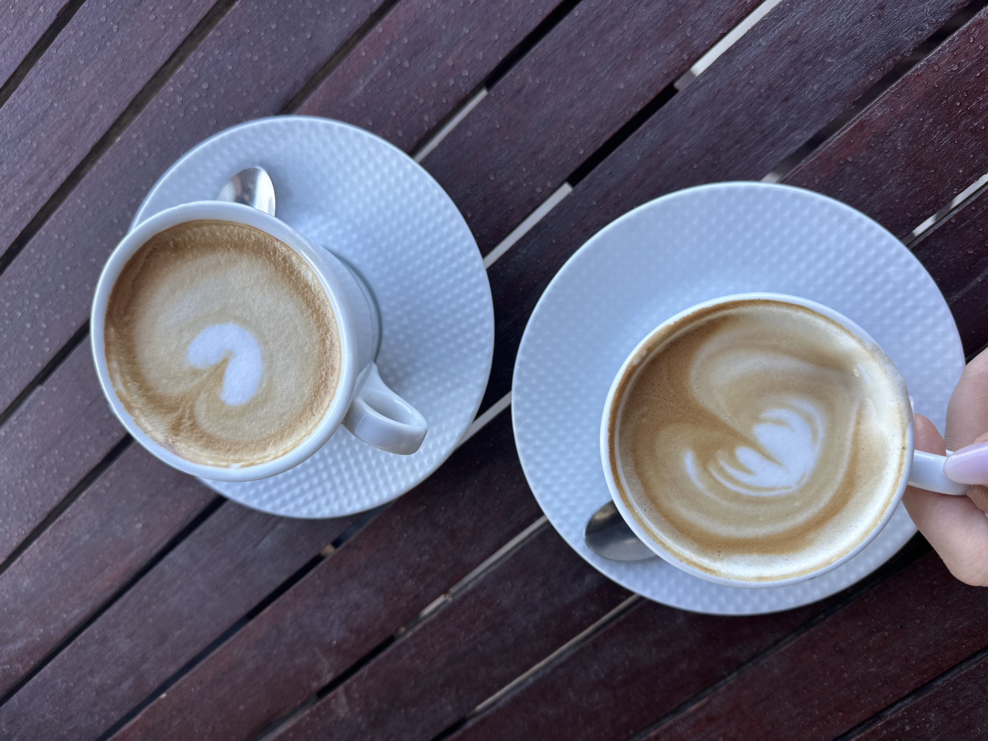 Two cups of coffee are placed on a wooden table. The coffee is served in white cups with white saucers and includes latte art. One cup is being held by a hand with a light pink nail polish. The location is likely outdoors given the wooden table, but the specific location in Šušanj, Montenegro is unidentifiable from the image.