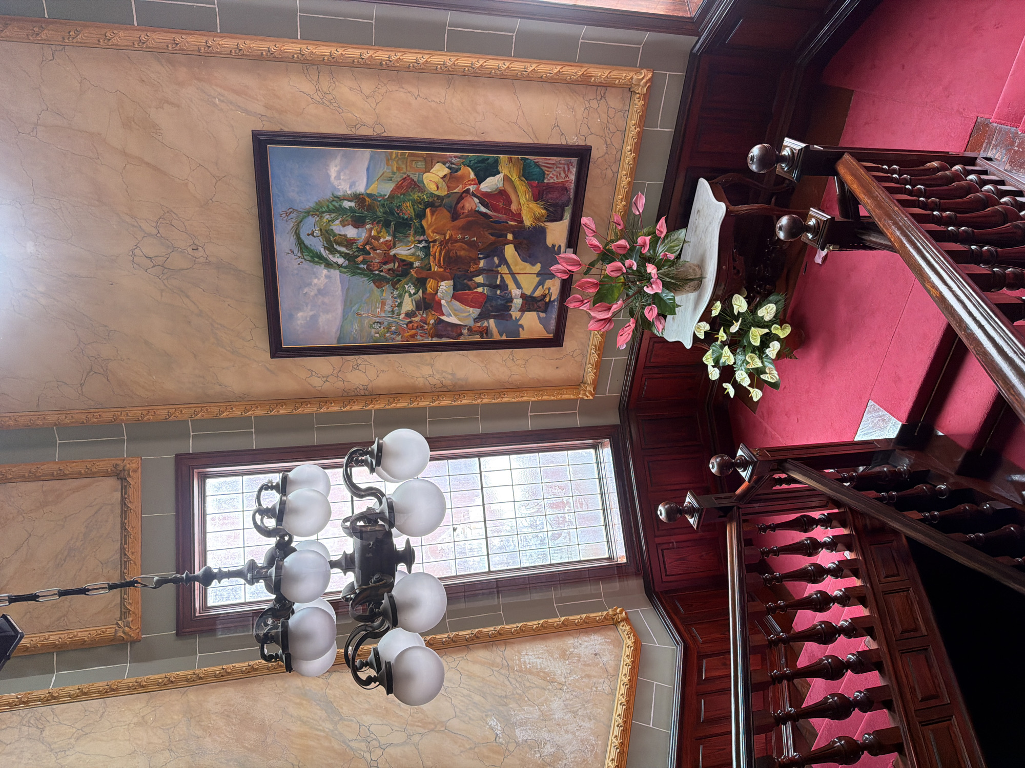 This is a high-angle interior shot of a grand staircase in La Orotava, Spain, likely within a historic building. The staircase features dark wooden banisters with intricately carved spindles and is covered in a vibrant red carpet. The walls are paneled with dark wood at the lower level, transitioning to a decorative marble-effect finish with ornate gold frames at the upper levels. A large framed painting depicting a festive scene with people and oxen is prominently displayed on the wall. Above the staircase, a multi-light chandelier with frosted globes hangs from the ceiling. A tall window with a grid pattern is visible behind the chandelier. On a marble-topped table near the bottom of the stairs, a vase filled with pink anthuriums and other greenery is present, with another potted plant of white anthuriums beside it. The scene appears to be well-maintained and opulent.