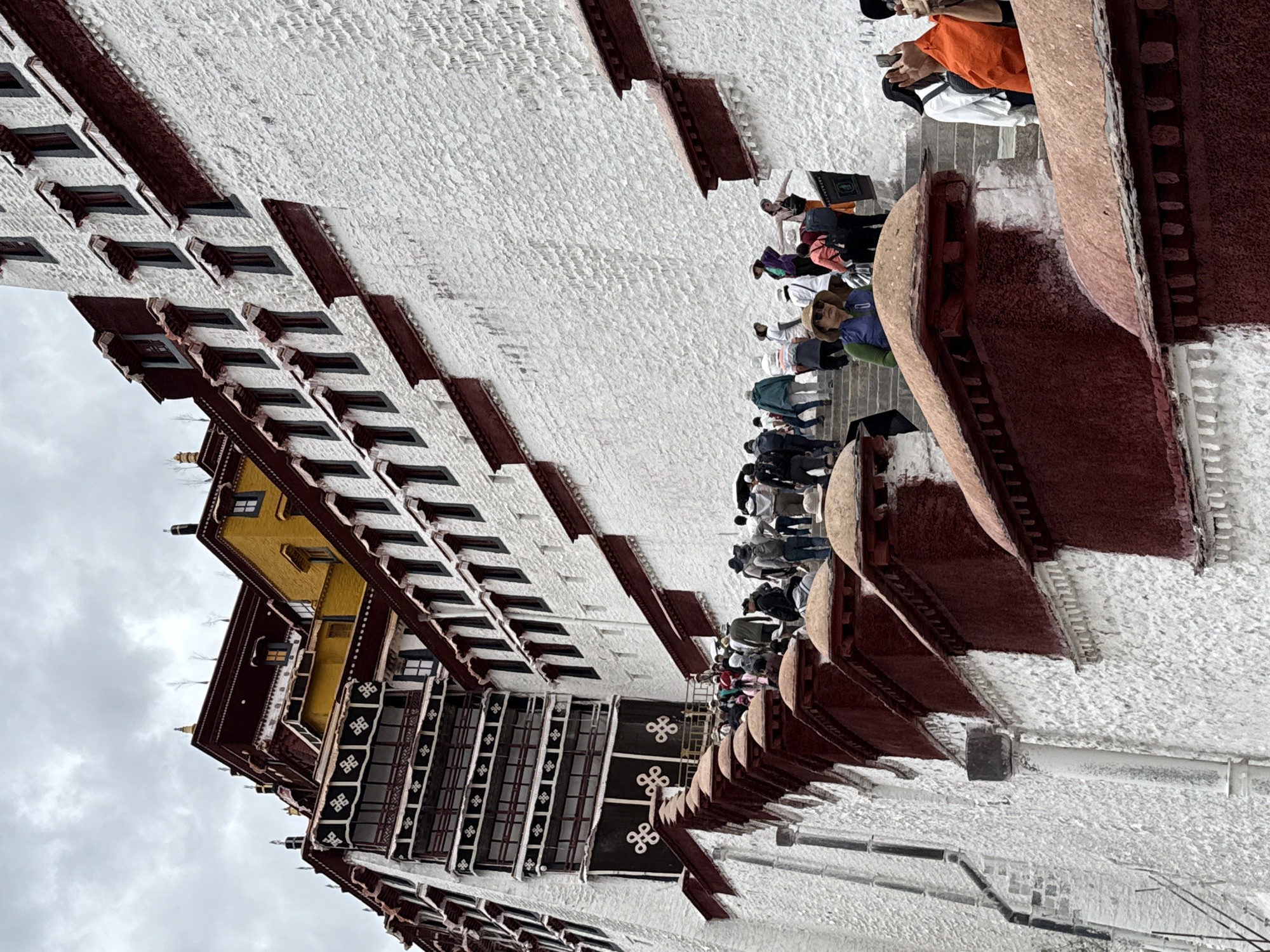 The image captures the Potala Palace in Lhasa, Chengguan District, China, a massive white-walled complex with red and gold accents. A crowd of people is visible on the steps leading up to the palace, appearing to be tourists. The palace has a distinct architecture, featuring numerous windows and protruding decorative elements. The sky is overcast.