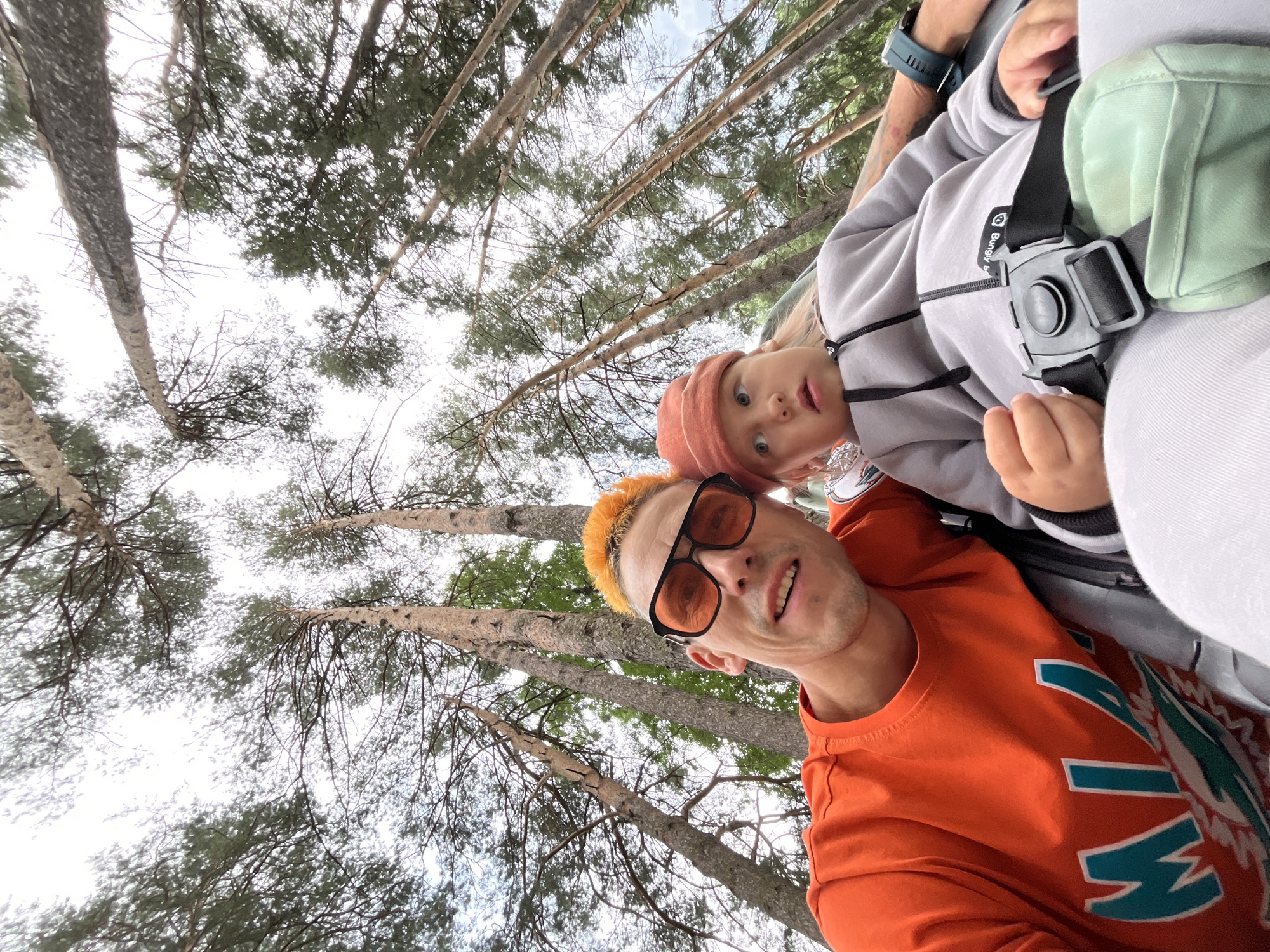 The image shows a low-angle, wide shot of two individuals, an adult male and a young child, looking up at the camera from what appears to be a seated position. The adult, on the left, has brightly dyed orange hair, is wearing sunglasses with orange lenses, and an orange t-shirt with the word "MIAMI" printed on it. The child, on the right, is wearing a coral-colored beanie and a gray hooded jacket. They are seated next to each other. The background consists of a dense forest of tall, slender pine trees reaching towards an overcast sky. The scene is outdoors, likely in a park or wooded area within the city of Chaikovsky, Russia, given the context of the prompt. The image seems to capture a casual moment between the two individuals during an outing in nature.