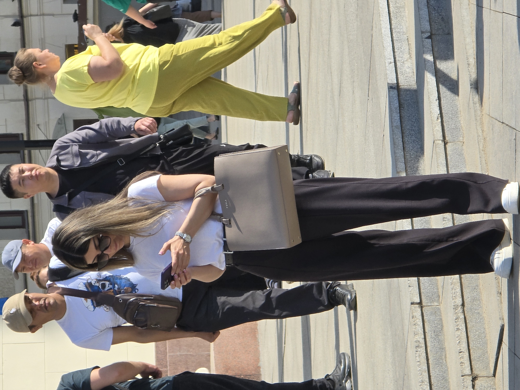 The image depicts several people walking on a city street. The setting is likely in Moscow, Russia, given the style of architecture and urban environment.
From top to bottom:
* A woman in a yellow top and pants is looking to the right.
* A man in a grey jacket and black shirt.
* A woman with long hair, sunglasses, a white t-shirt, and black pants is holding a phone and carrying a grey purse.
* Two other men are wearing baseball caps and casual shirts.
* The bottom portion of the photo shows legs and shoes of another person wearing dark clothing.
The street has stone or tile pavement.