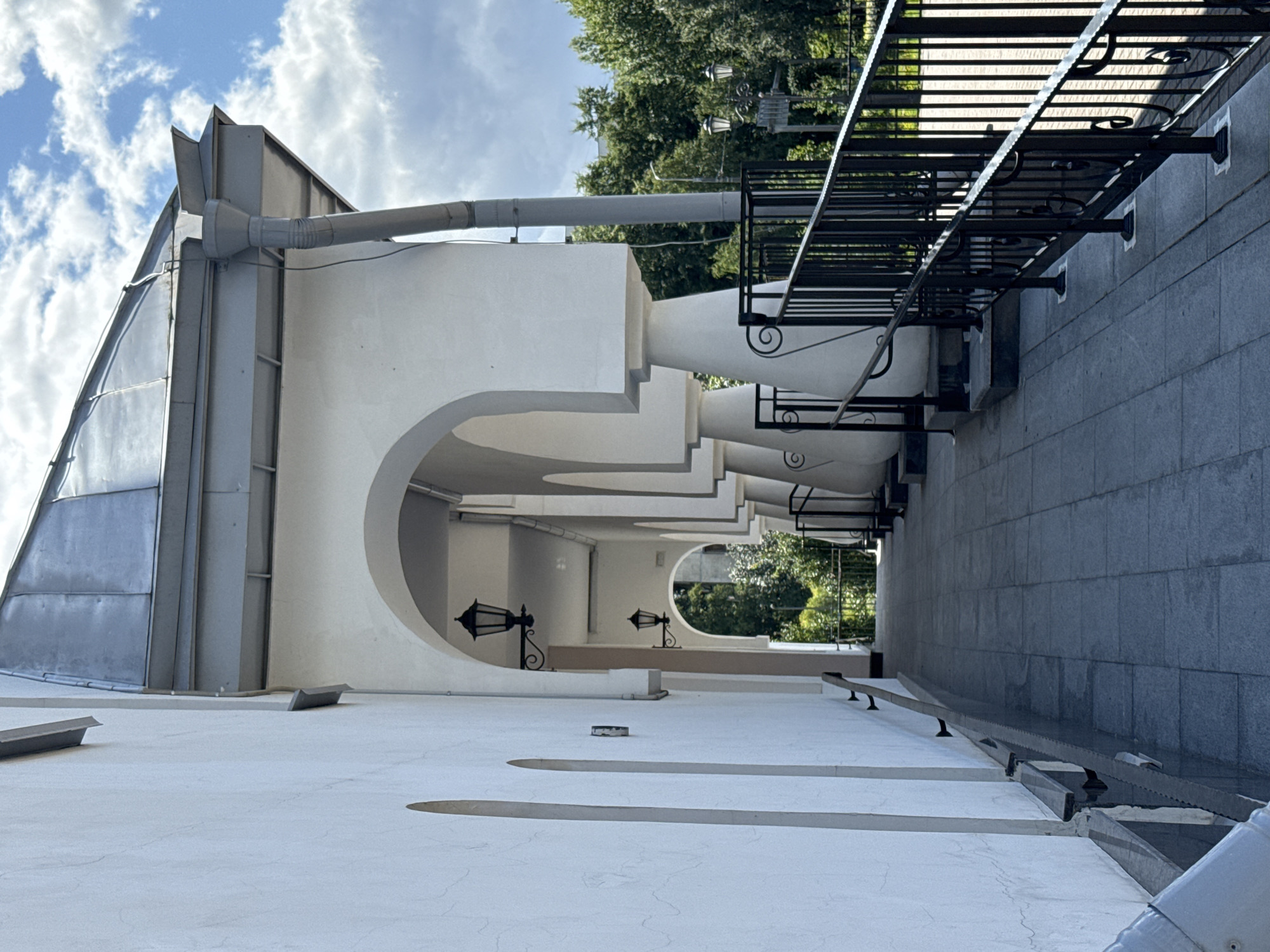 The image shows a covered walkway with a series of arched openings, leading into the distance. The walkway is paved with square gray tiles. To the left, a plain white wall features two narrow, recessed arched niches. On the right side of the walkway, a black metal railing with decorative scrollwork follows the path. Behind the railing, large white decorative columns support the roof of the walkway. Two black ornate lanterns are visible mounted on the wall inside the arched openings. Beyond the walkway, trees and greenery are visible, suggesting an outdoor setting. The sky above is blue with scattered white clouds. The overall scene depicts an architectural element of a building, possibly a historical one. The context provided indicates the location is Saint Petersburg, Russia.