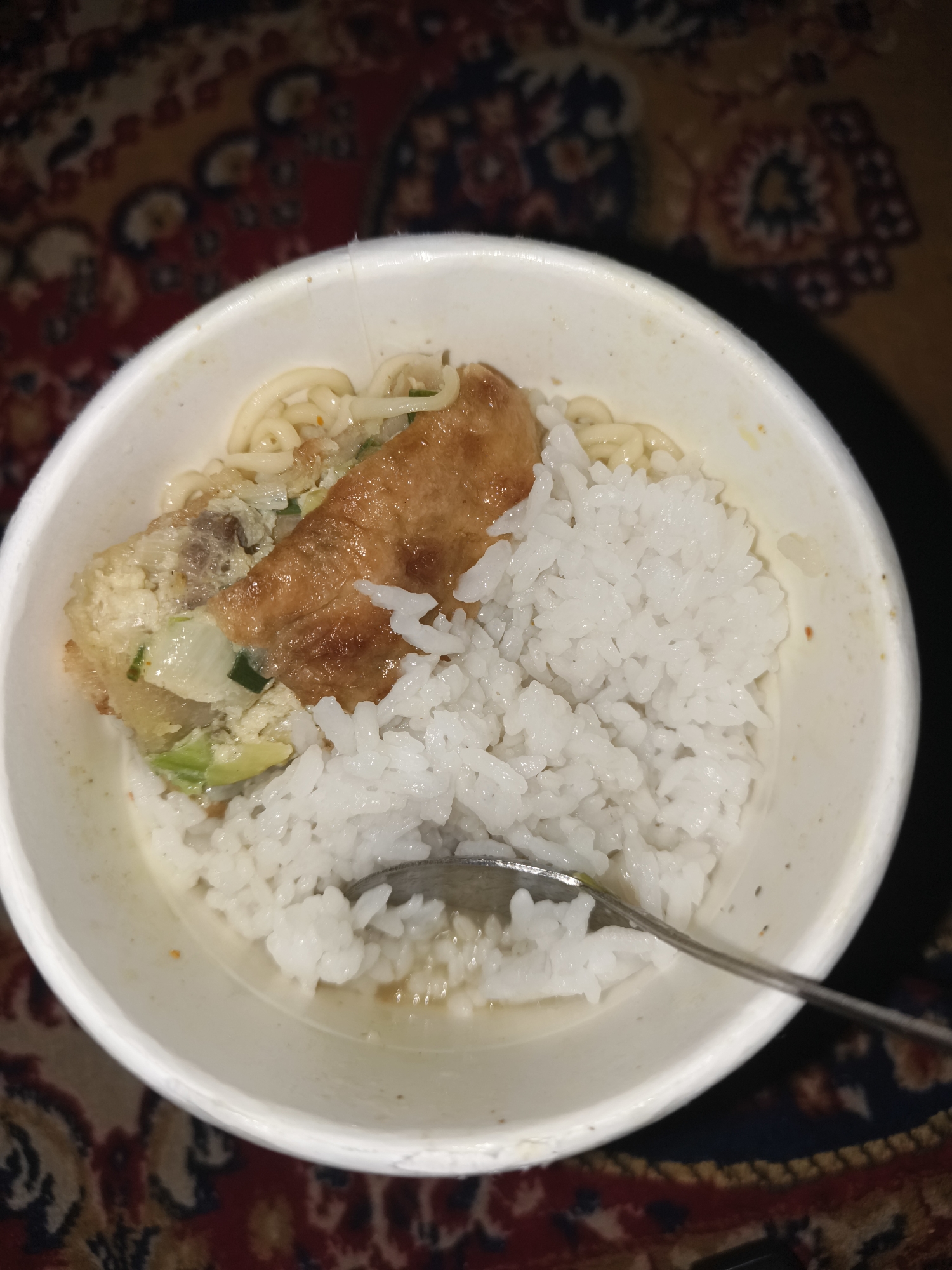 A close-up, top-down view shows a white, disposable bowl filled with food. The bowl contains a portion of white rice to the right and what appears to be a fried piece of food with some visible green vegetables and noodles beneath it, all immersed in a light-colored broth. A silver spoon is partially visible in the bottom right corner, resting in the broth near the rice. The bowl is placed on a surface with a patterned, reddish-brown and yellow textile. The overall lighting is dim, with some artificial light source creating reflections on the broth and spoon. The location in the city of Barengkrajan, Indonesia, is not discernible from the image.