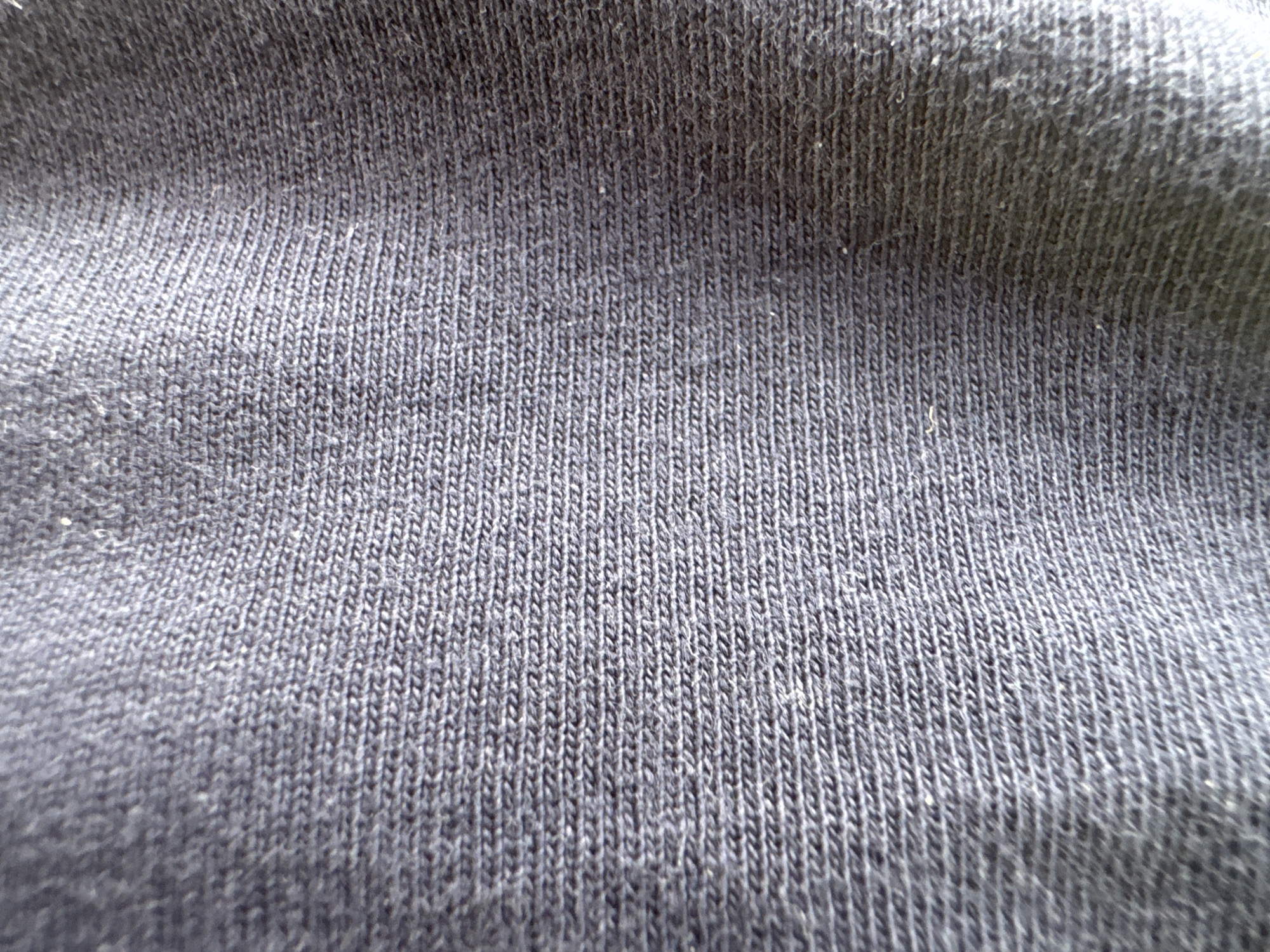 The image is a close-up, high-angle shot of a dark navy blue knitted fabric. The texture of the knit is clearly visible, with a fine ribbing pattern running horizontally across the frame. The fabric appears soft and slightly fuzzy, with some subtle variations in shade due to the way light reflects off the fibers. There are no people, other objects, or environmental details visible, making it an abstract textural study. The location in Strümpfelbrunn, Germany is not discernible from the image content.