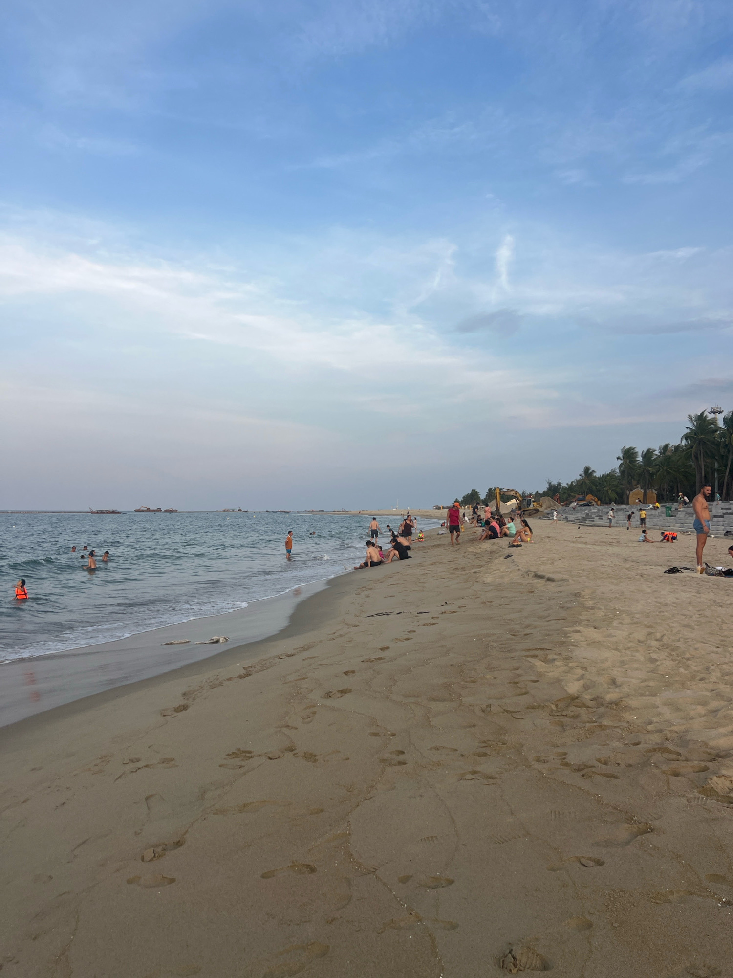 This is a medium-shot, eye-level, outdoor, vertical, full-color photograph of a crowded beach in Hoi An, Vietnam. The foreground is dominated by the sandy beach with numerous footprints. The middle ground shows a beach with many people, some sitting and some standing. The sea is in the background on the left, with a few people swimming. The sky is light blue with scattered white clouds. Palm trees and construction equipment are visible in the distance on the right.