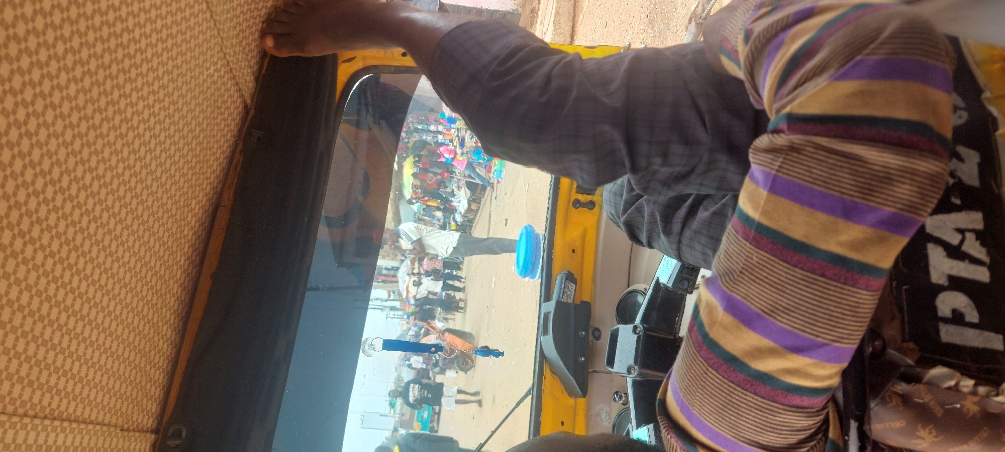 The image depicts a street scene viewed from the interior of a vehicle, likely a motorized rickshaw, with two occupants partially visible. The ceiling of the vehicle is covered in a light brown fabric with a repeating small checkered pattern. A dark frame separates the patterned ceiling from the windshield. Hanging from the windshield via a suction cup is a blue lanyard with a small, blue fish-shaped charm.
In the lower left, the left arm and shoulder of a person, presumably the driver, are visible, wearing a long-sleeved shirt with horizontal stripes in gold, purple, and green. Below this arm, part of a black surface with the white text "PTA" is discernible. In the upper right, the bare right foot and lower leg of another person are visible, propped against the vehicle's frame. This person is wearing dark, possibly plaid or checkered trousers. The vehicle's interior features a yellow-painted metal frame and parts of the dashboard/steering mechanism.
Outside the vehicle, a bustling street or marketplace is visible under an overcast sky. The ground is unpaved and appears sandy or dusty. Numerous people are present. A man in a white shirt with dark sleeve accents and dark trousers stands prominently in the center-right, facing left. A blue plastic basin or bowl rests on the ground near his feet. Along the right side of the street, market stalls display various colorful goods, possibly plastic items, under temporary coverings. Further to the left, a light blue three-wheeled vehicle is partially visible. In the background, a multi-story building and utility poles are faintly visible, indicating an urban setting in Unknown, Unknown.