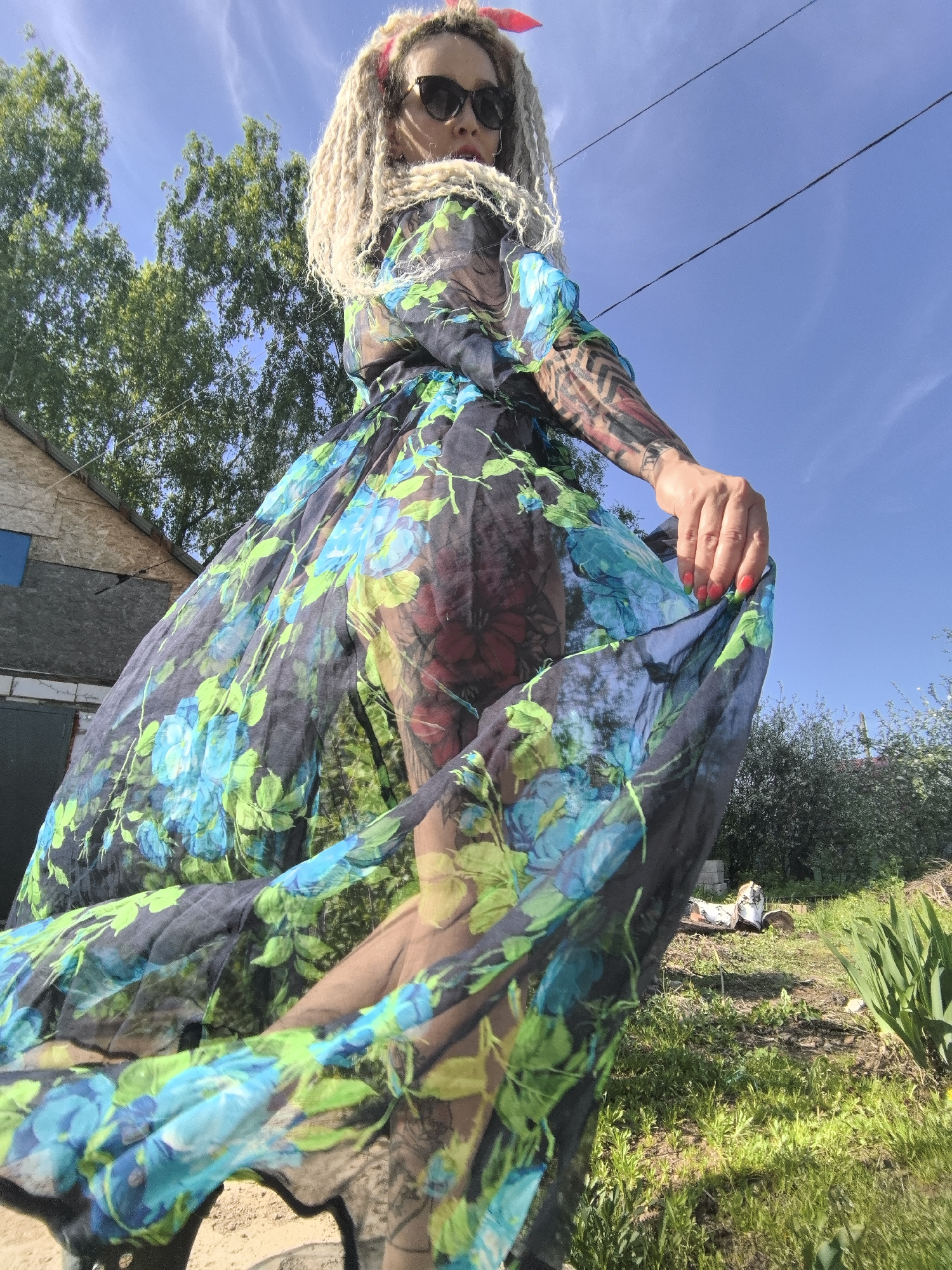 A woman with blonde dreadlocks, sunglasses, and a red hair accessory is seen from a low angle in Noviy, Russia. She wears a flowing, translucent dress with a dark background and large blue and green floral patterns. The dress is partially obscuring a tattoo on her arm and leg. She is holding the edge of the dress, causing it to billow.
In the background, a small, weathered building with a dark green door is visible to the left. The sky is blue with scattered clouds. Green grass, foliage, and miscellaneous objects like logs and debris are visible in the yard. Power lines are also visible in the sky.