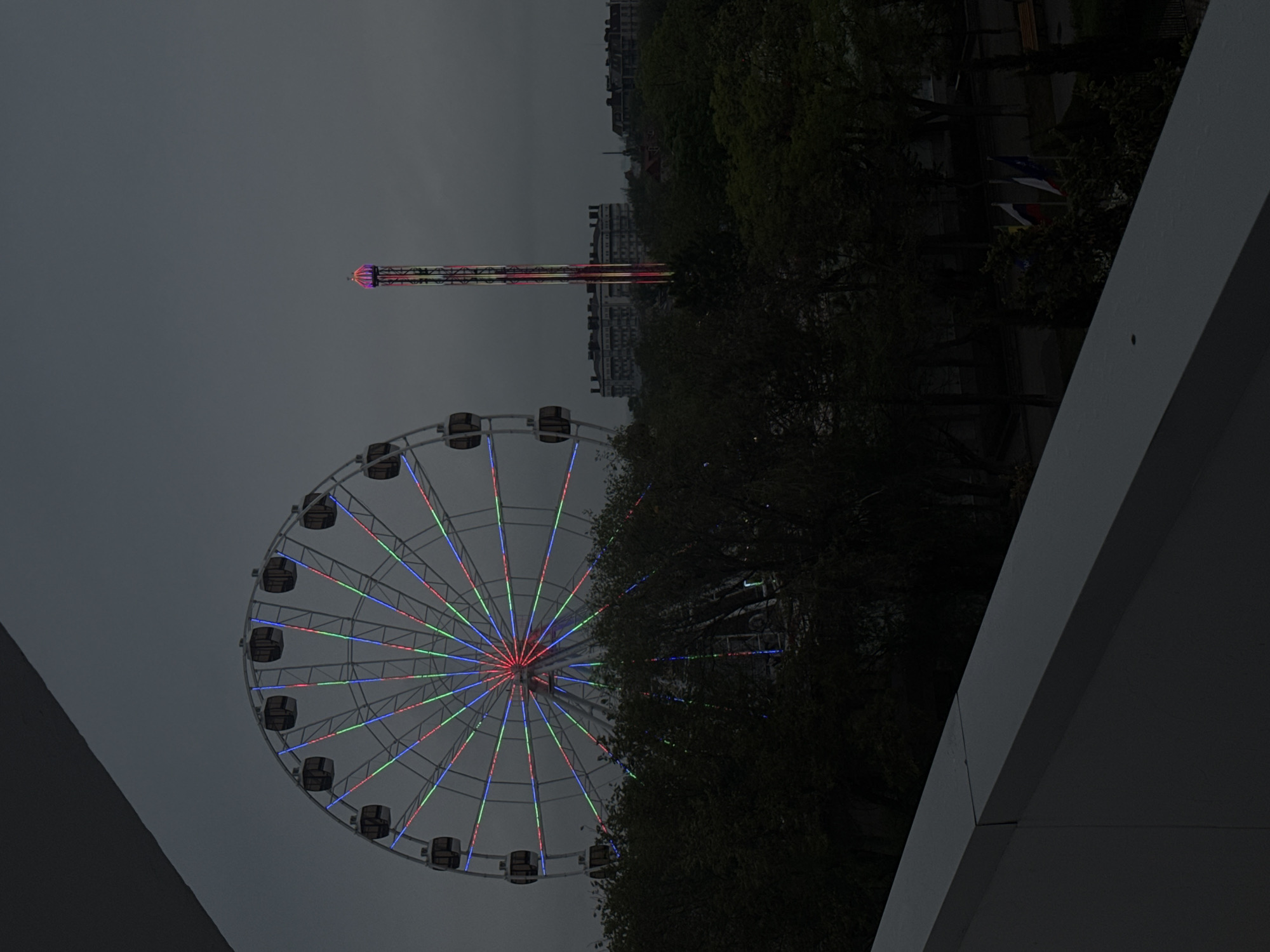 The image depicts a Ferris wheel and a tall amusement park ride in Yevpatoriya, Ukraine. The Ferris wheel is adorned with multicolored lights. A tall structure, also illuminated, stands nearby. The scene is viewed through the framing of a modern architectural structure. Buildings and trees are visible in the background, contributing to the urban setting. The sky is overcast.