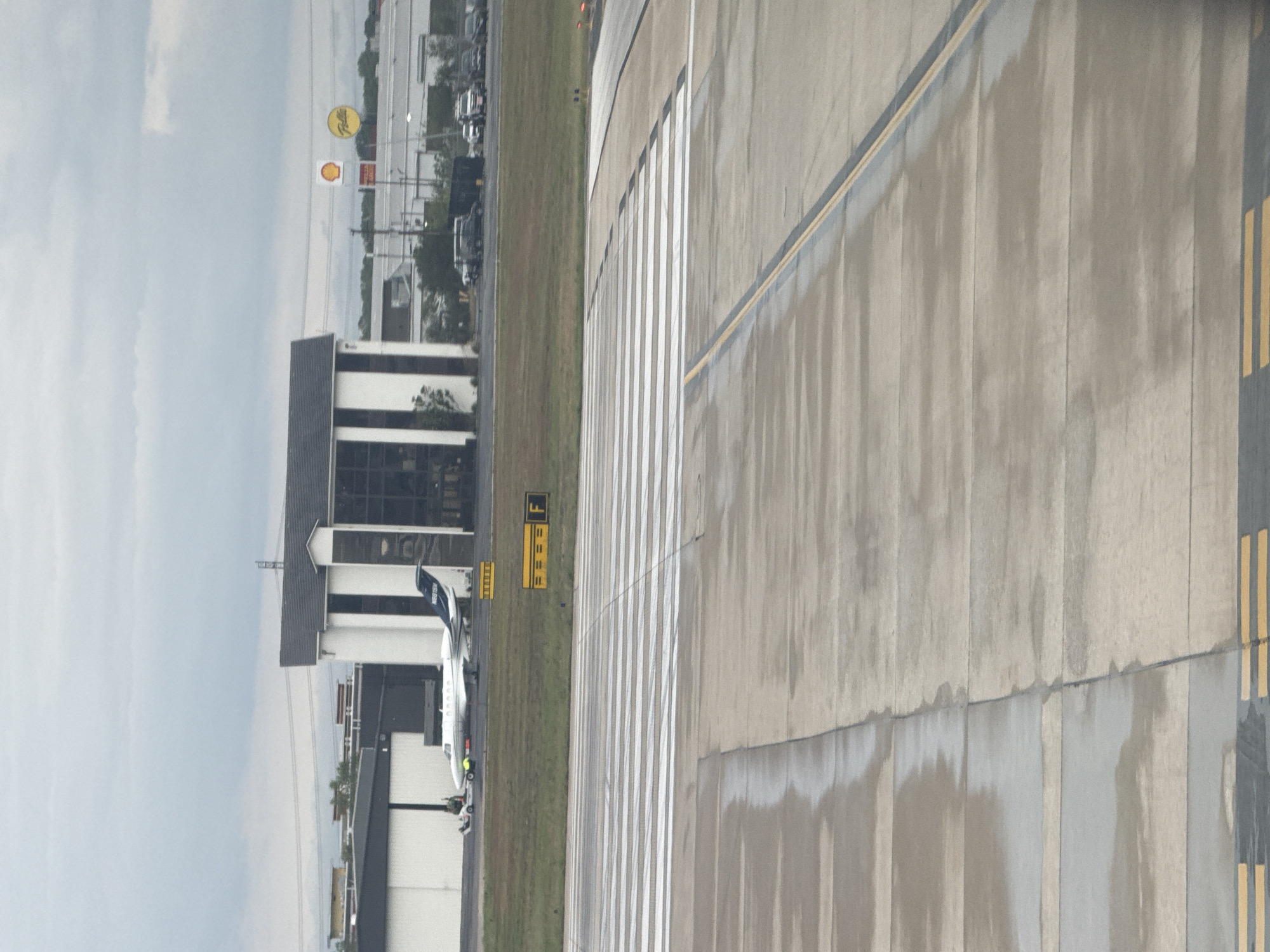 The image shows a view from what appears to be an airport tarmac. The location appears to be the San Antonio International Airport, indicated by the Shell and Pilot gas station signs. A small jet is parked at a building adjacent to the tarmac. The tarmac is marked with white and yellow lines, as well as painted designations. The weather is overcast.