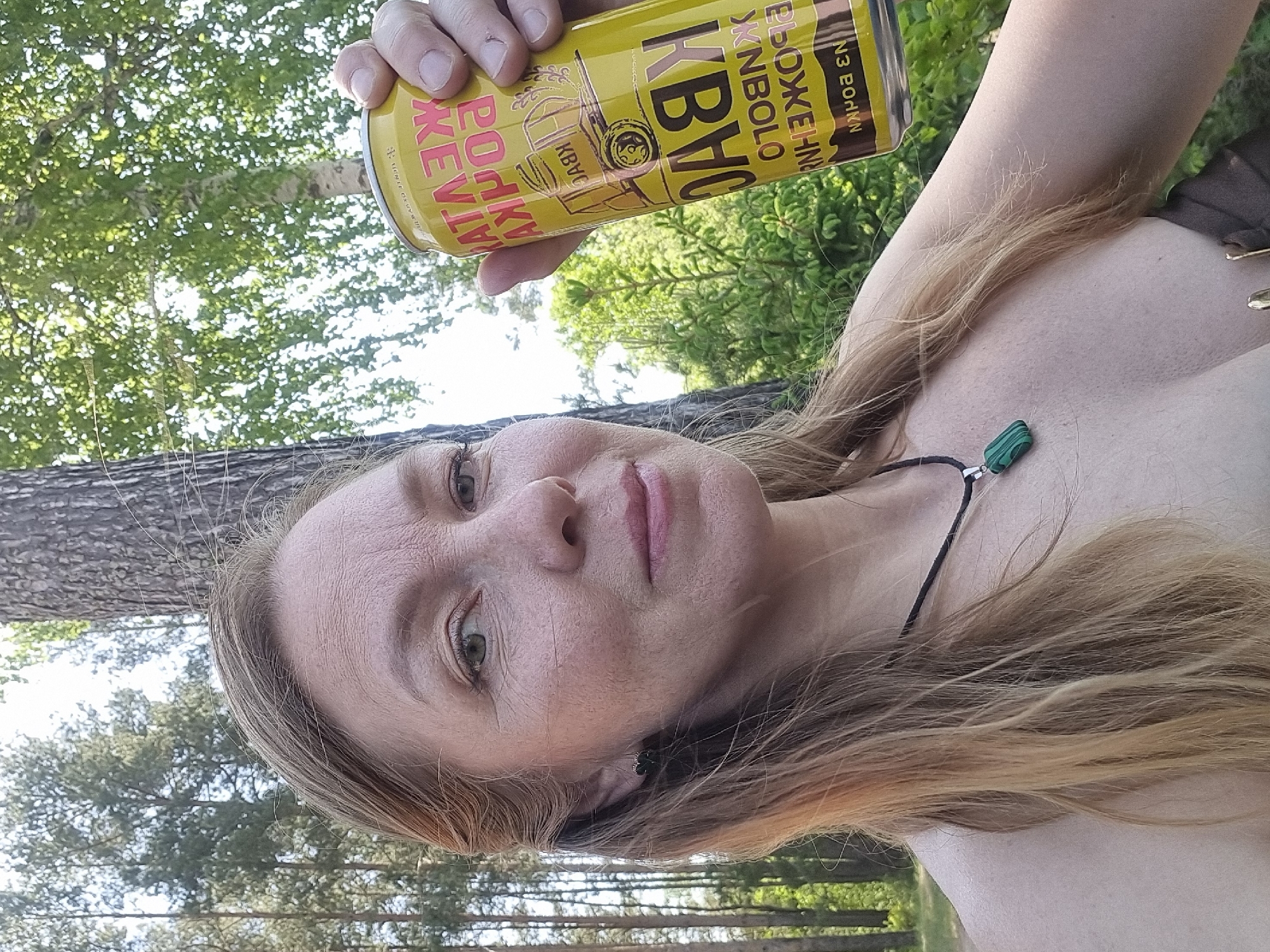The image is a selfie taken outdoors in Chaikovsky, Russia. A fair-skinned woman with light brown hair is holding a yellow can of "Kvas" brand beverage. The woman is wearing a black cord necklace with a green stone pendant and a small earring. The background shows a forest with trees visible. The photo appears to have been taken during daylight hours.