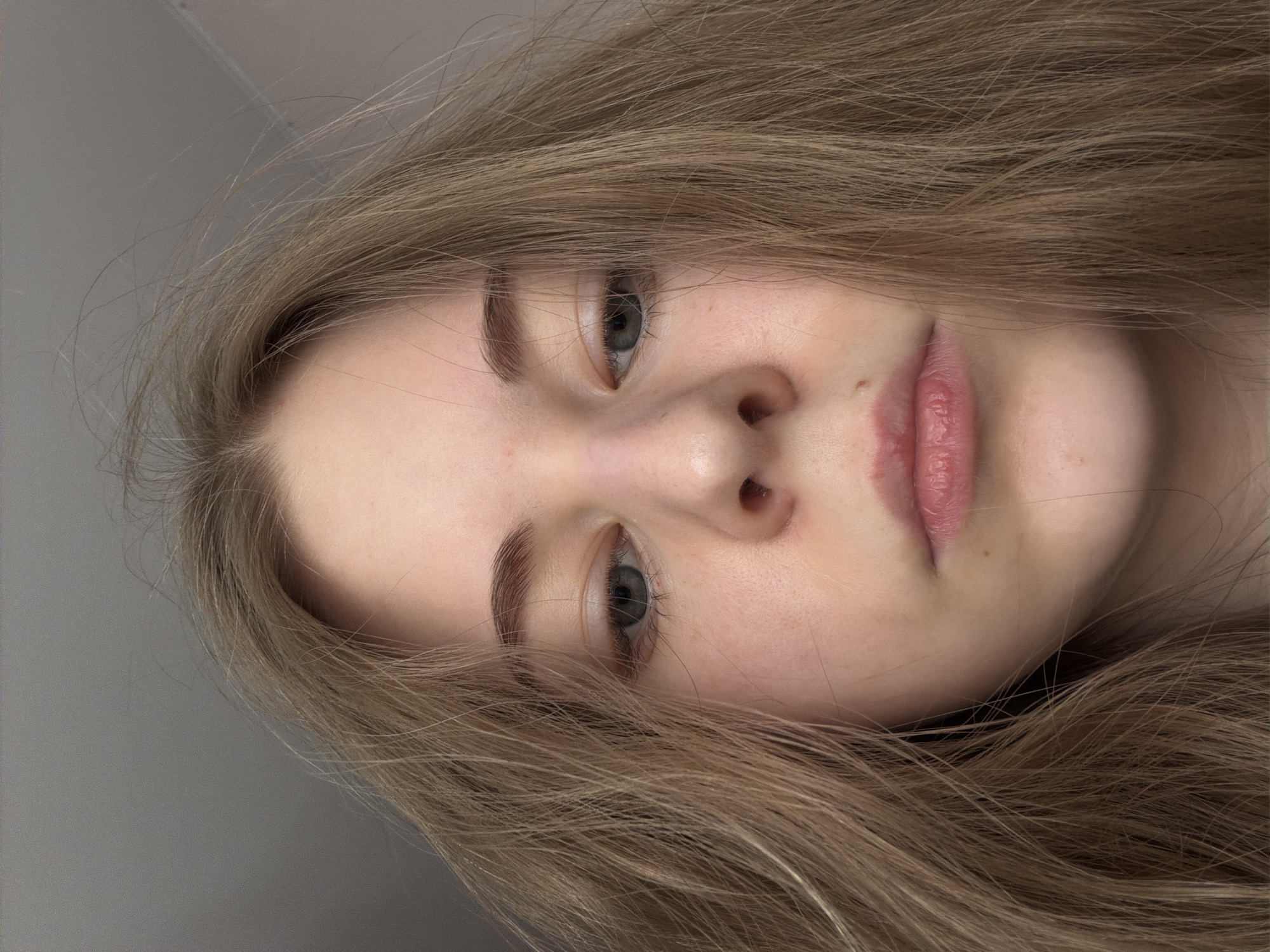 A close-up, slightly rotated image depicts a young woman with fair skin, light brown hair, and light-colored eyes. Her face is centered, occupying the majority of the frame. Her lips are pink and slightly parted. The background is a solid gray color. The photograph provides no indication of the city of Perm, Russia or any other specific location.