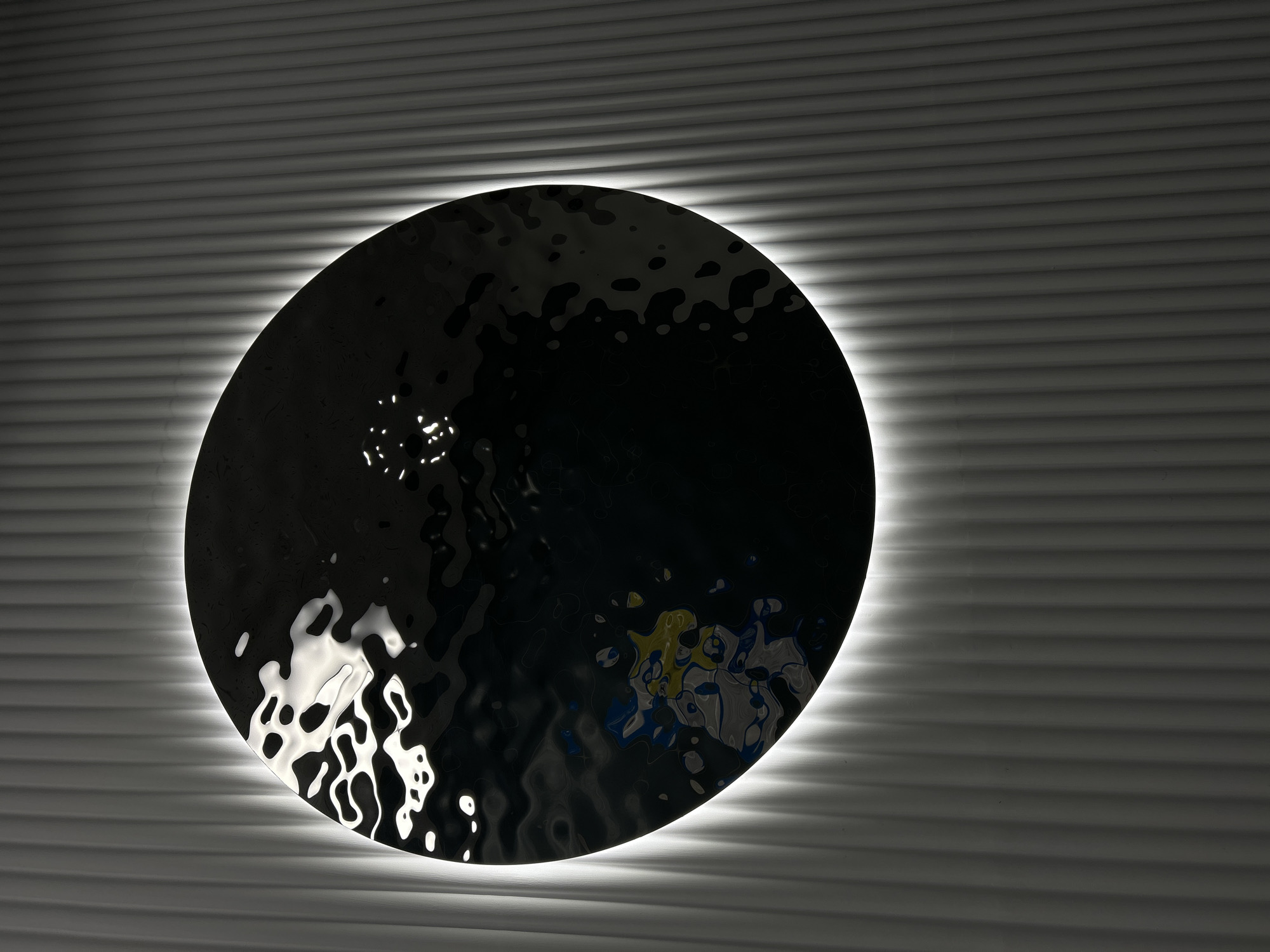 A dark, circular, hammered-metal sculpture is mounted on a horizontally ribbed wall. A soft, white light emanates from behind the sculpture, casting shadows on the wall. Reflections of other colours are visible on the surface of the sculpture. The setting appears to be indoors. There is no information in the provided images that can confirm this photo was taken in Saint Petersburg, Russia.