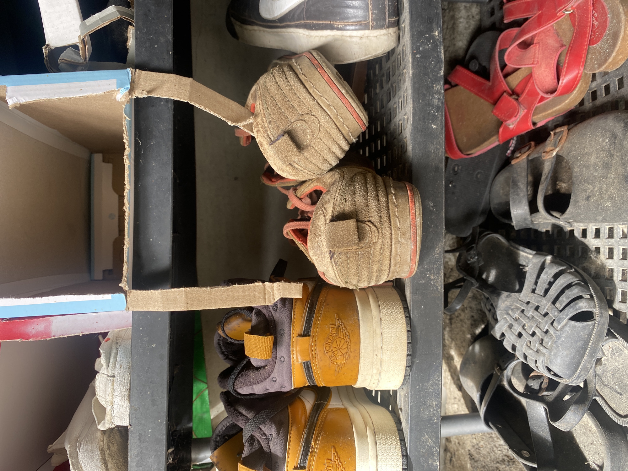 This image captures a close-up view of various footwear stored on and around a multi-tiered shelving unit, likely in a household setting in Davao City, Philippines. The scene is illuminated by natural daylight, creating some shadows.
The primary subjects are numerous pairs of shoes and sandals, most of which show signs of wear and dust. On the top left, a dark brown shoe with a white Nike swoosh is partially visible. Below it, two small, beige or light brown children's shoes with orange trim and laces are placed, appearing to be soft-soled.
To the right, a vibrant pair of red sandals are stacked, along with several darker-colored sandals, including black ones with buckled straps and a woven design. These are resting on a dark, grid-patterned surface. At the bottom left, two pairs of brown and black high-top sneakers are prominent, featuring the "AIR JORDAN" logo embossed on their sides. Another light-colored shoe is partially seen beneath these.
The footwear is arranged within a storage system composed of a dark, possibly metal, frame. On the left side, an improvised shelving or divider structure made from cardboard, showing blue and white markings and some wear, is attached to the main frame. The overall impression is a practical, somewhat cluttered storage solution for a family's shoes. No human activity or specific event is taking place; it is a static display of stored items.