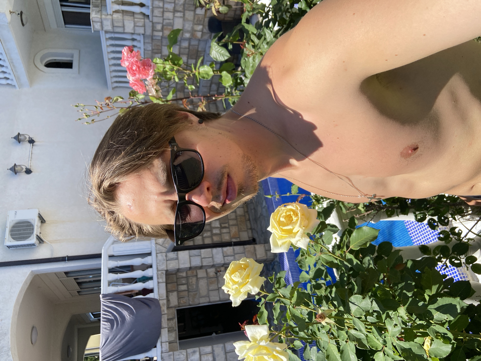 A medium shot shows a shirtless man with sunglasses near roses and building facade outdoors. The man has light brown hair, facial hair, and a silver necklace. He is near a rose bush with yellow and pink roses. There is a white two-story building with a stone and stucco facade and air conditioning unit on the side. The image was taken in Šušanj, Montenegro.
