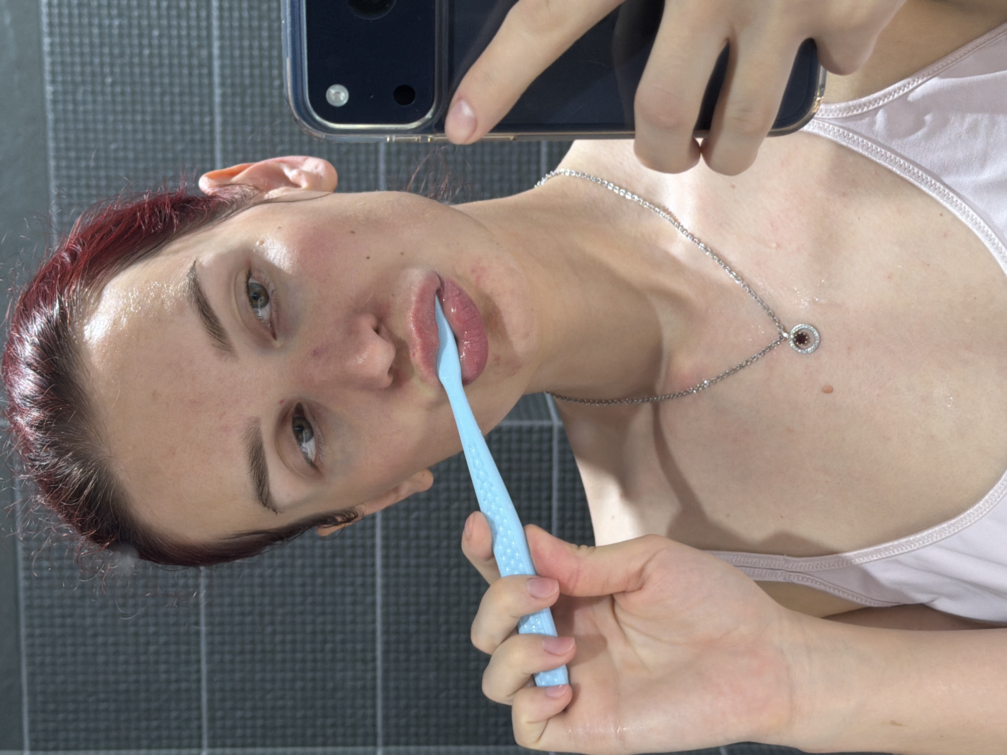 A young woman is taking a selfie while brushing her teeth in a bathroom. She is holding a smartphone in her right hand, partially obscuring the upper portion of the image. She is wearing a light-colored tank top and a silver necklace with a circular pendant. Her hair is dark red. A light blue toothbrush is in her left hand. The background shows gray tiled walls. No information is available to determine the exact location of the bathroom, and there is no visual information in the image to confirm that the location is Chaikovsky, Russia.