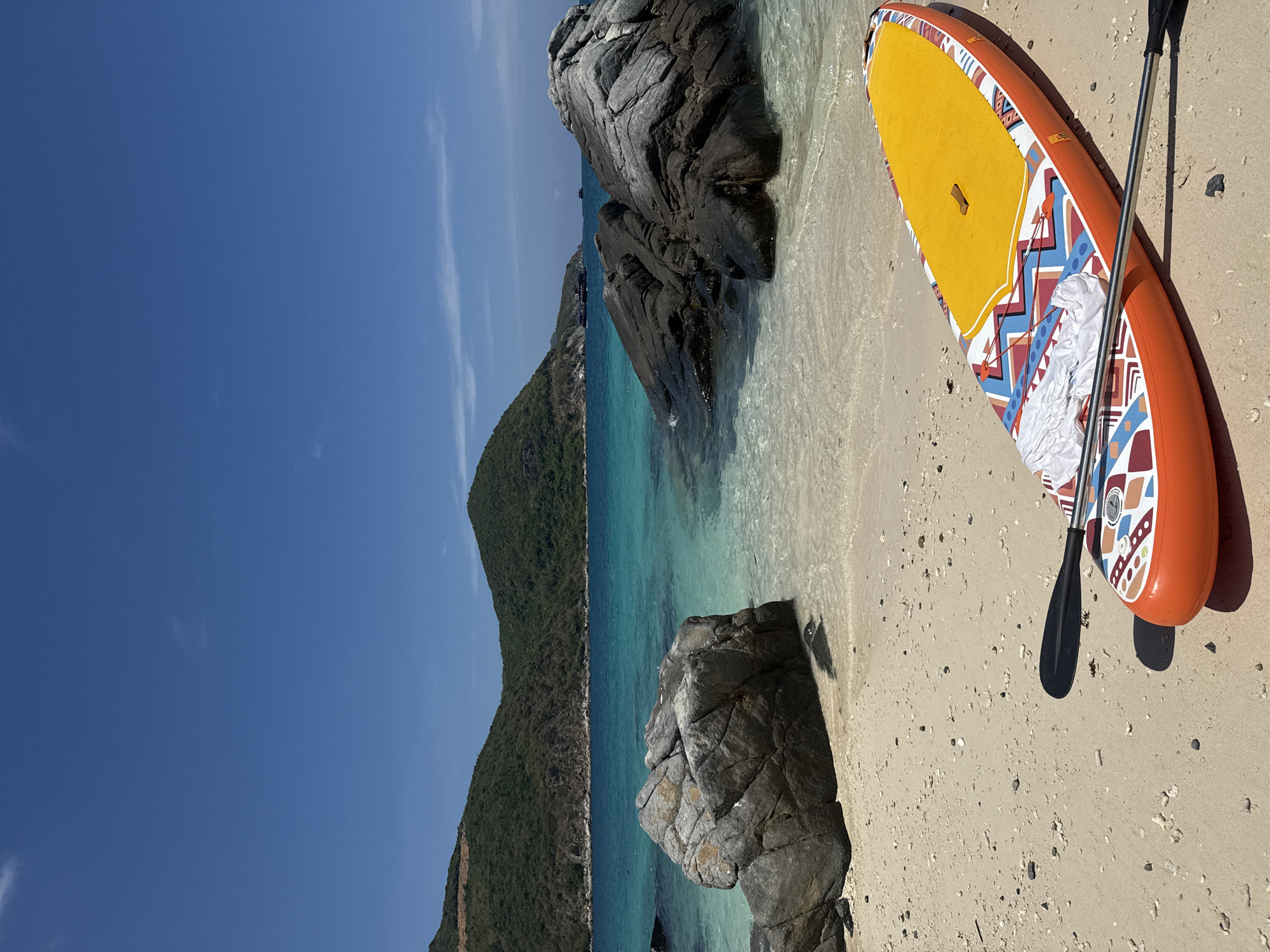 The image shows a beach scene in Nha Trang Ward, Vietnam. A colorful stand-up paddleboard with a paddle rests on the sandy beach near the water's edge. Large rocks are scattered along the shoreline. A green, forested hill rises in the background, and the sea is a clear turquoise color. The sky is a clear blue.