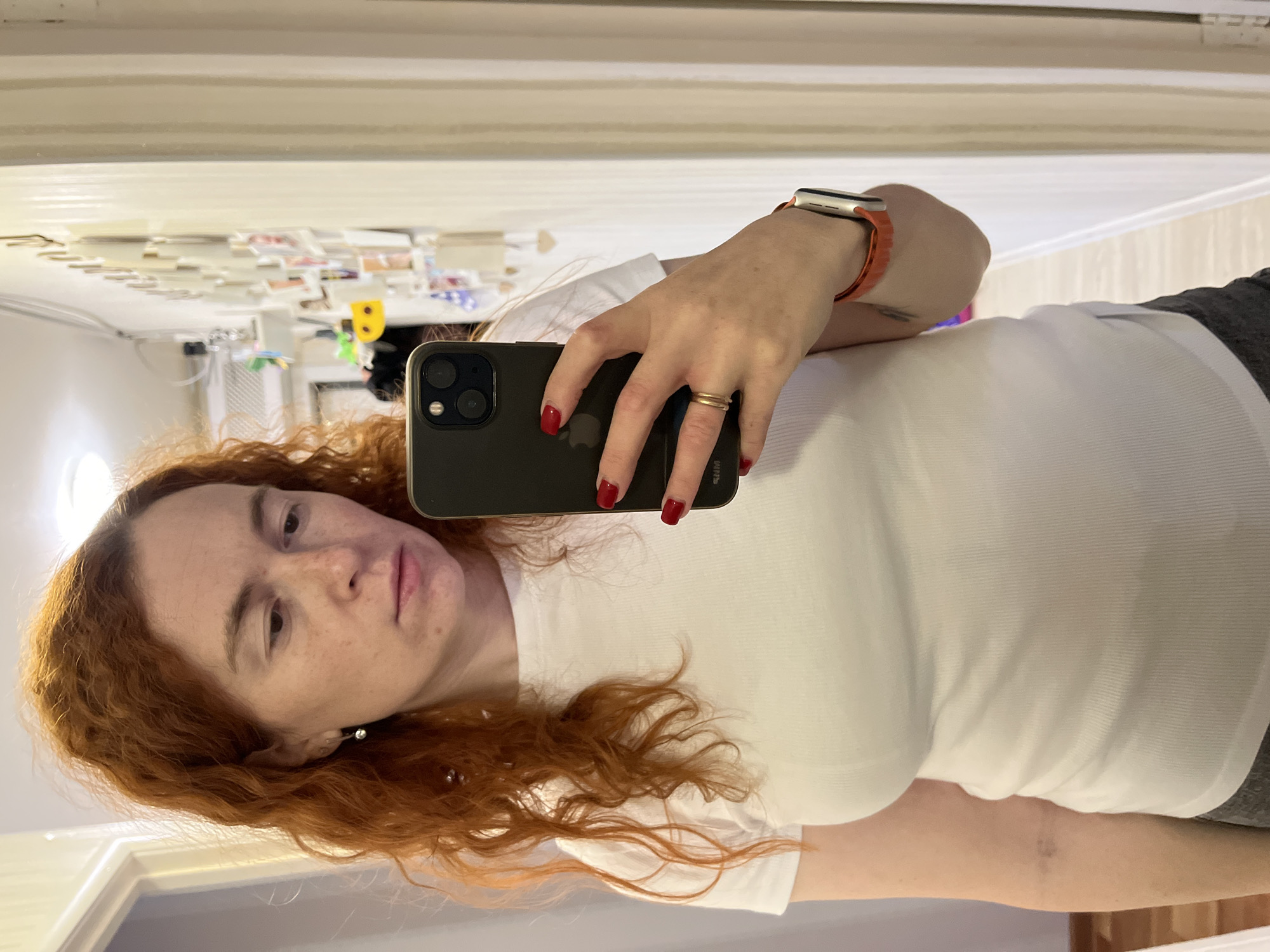 A woman is taking a selfie in what appears to be an indoor setting. She has red hair and is wearing a white t-shirt, an Apple Watch with an orange band, and a ring on her left ring finger. She's holding a dark-colored iPhone in her hand. The background includes a mirror, which reflects part of a room, but there is no indication of the location being Chaikovsky, Russia.