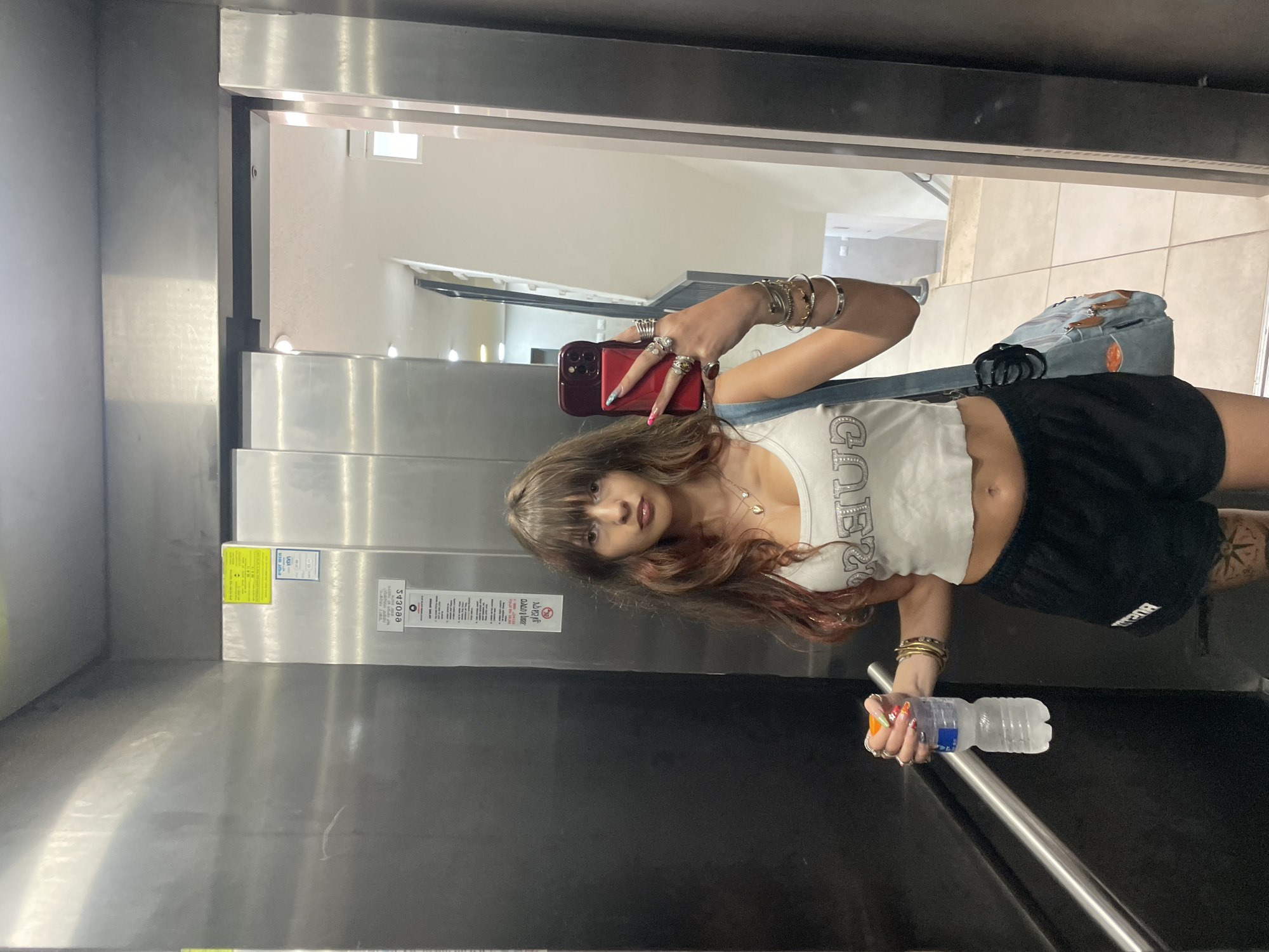 A young woman with long, wavy brown hair with streaks of red and blonde stands in an elevator, taking a selfie. She is wearing a white, cropped tank top with the word "GUESS" in silver lettering, and black athletic shorts. She has a collection of silver bracelets on her left wrist and several rings on her fingers. In her left hand, she holds a clear plastic bottle of water. A light blue denim bag with tan leather accents hangs from her right shoulder. The elevator walls are made of brushed metal, and a metal handrail runs along the left side. A sticker with Hebrew text is visible on the elevator door. The floor is tiled. The scene appears to be a residential or commercial building in Raanana, Israel.