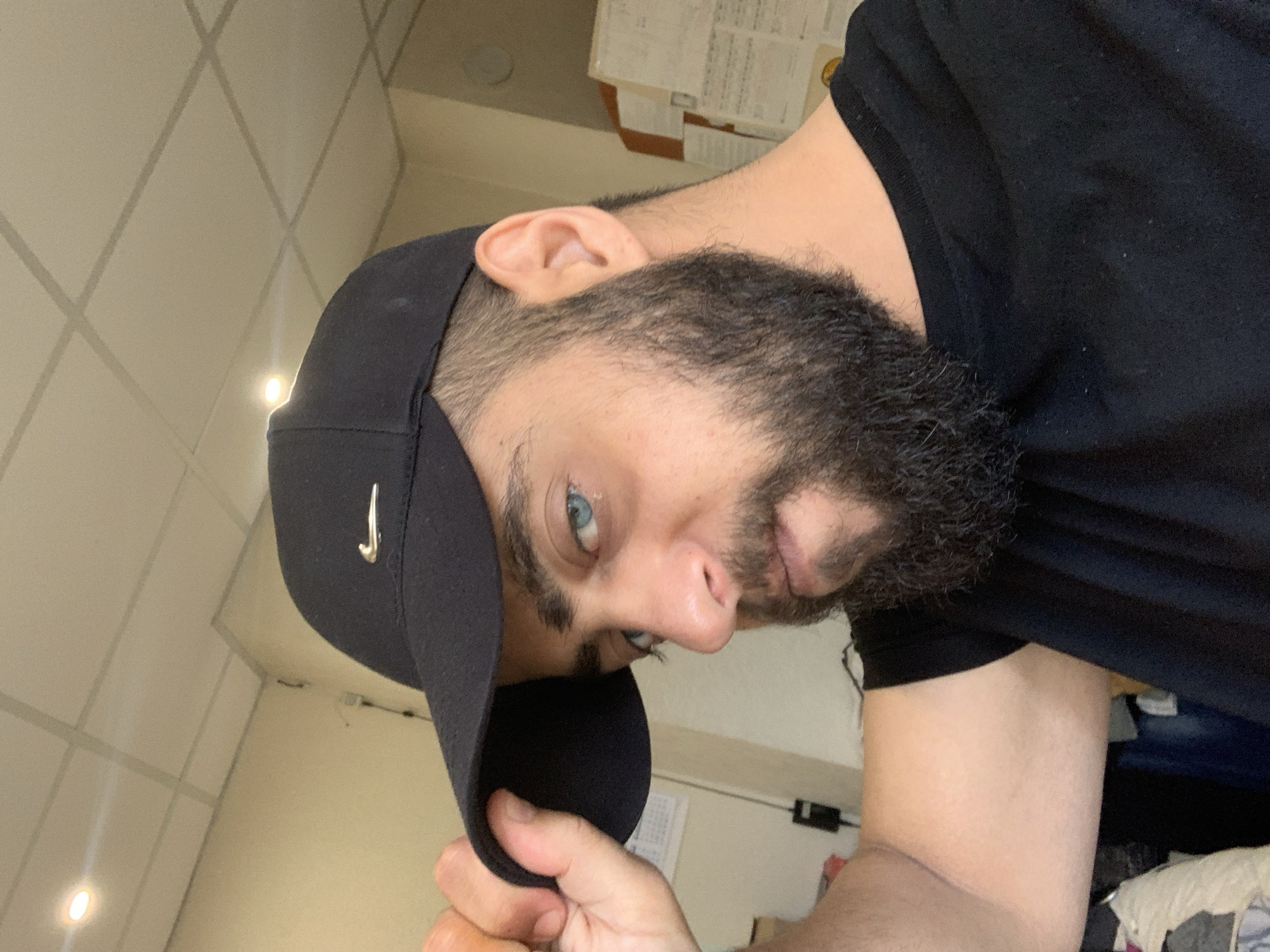 The image is a rotated, close-up indoor portrait of a man, taken from a low angle. The man has a dark, full beard and striking light blue eyes. He is wearing a black baseball cap with a silver logo, likely a Nike swoosh, on its side, and a black t-shirt. His left hand is visible, holding the brim of his cap. The man looks directly at the camera with a neutral expression. The background shows light-colored walls and a ceiling with a grid pattern, possibly acoustic tiles, and rectangular light fixtures. Stacked papers or documents are partially visible in the upper right. The image is rotated 90 degrees counter-clockwise, presenting the man's head horizontally.