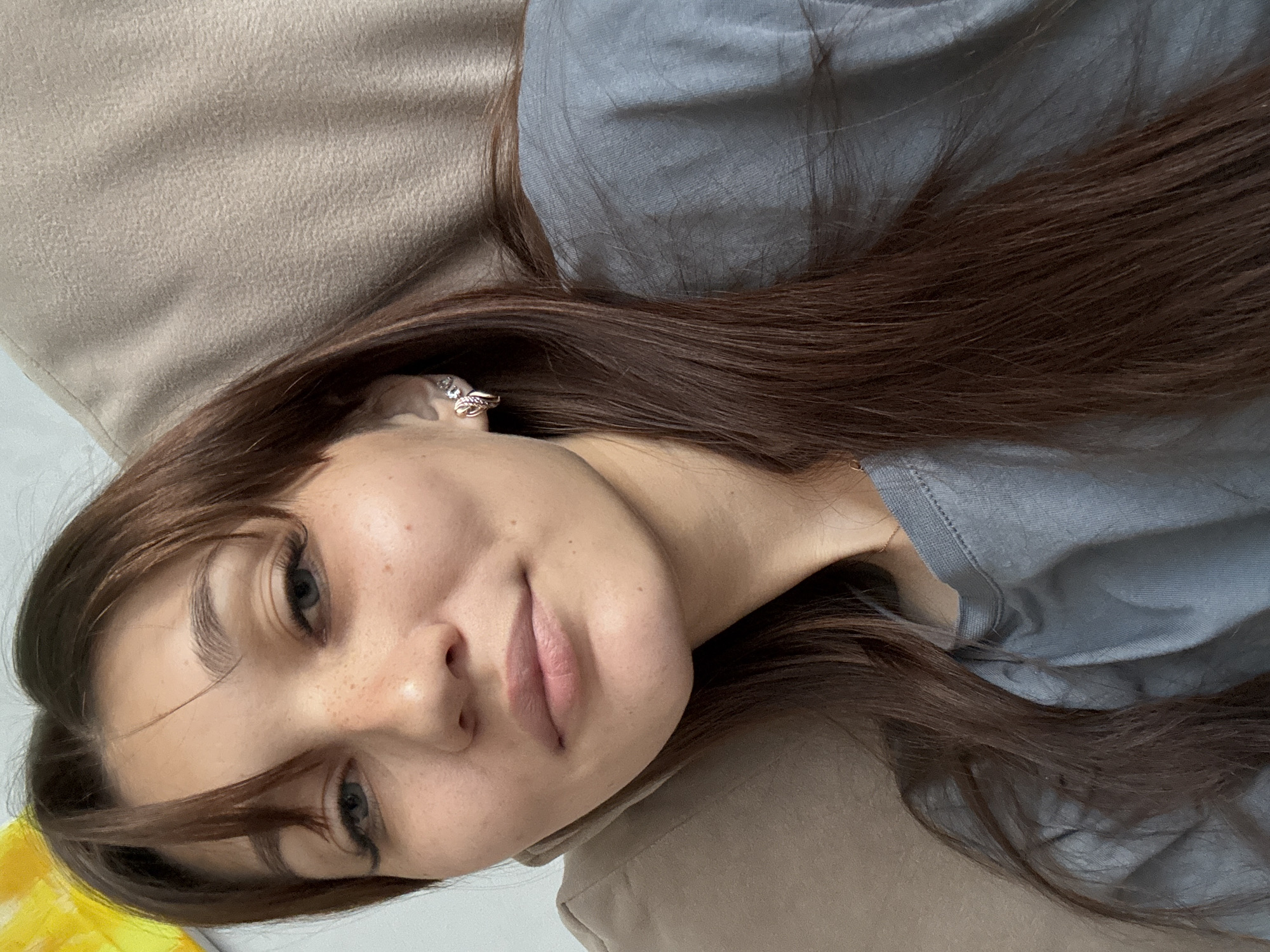 A young woman with long, dark brown hair and a gray t-shirt is shown from the chest up in a close-up, selfie-style photograph. She is wearing earrings and has freckles on her face. The background is a soft, beige surface, possibly a sofa or pillow, and a hint of a white wall is visible on the left. The lighting is soft and diffused. There are no other people or objects of note in the image. The location in Novokuznetsk, Russia, is not discernible from the visual information provided.