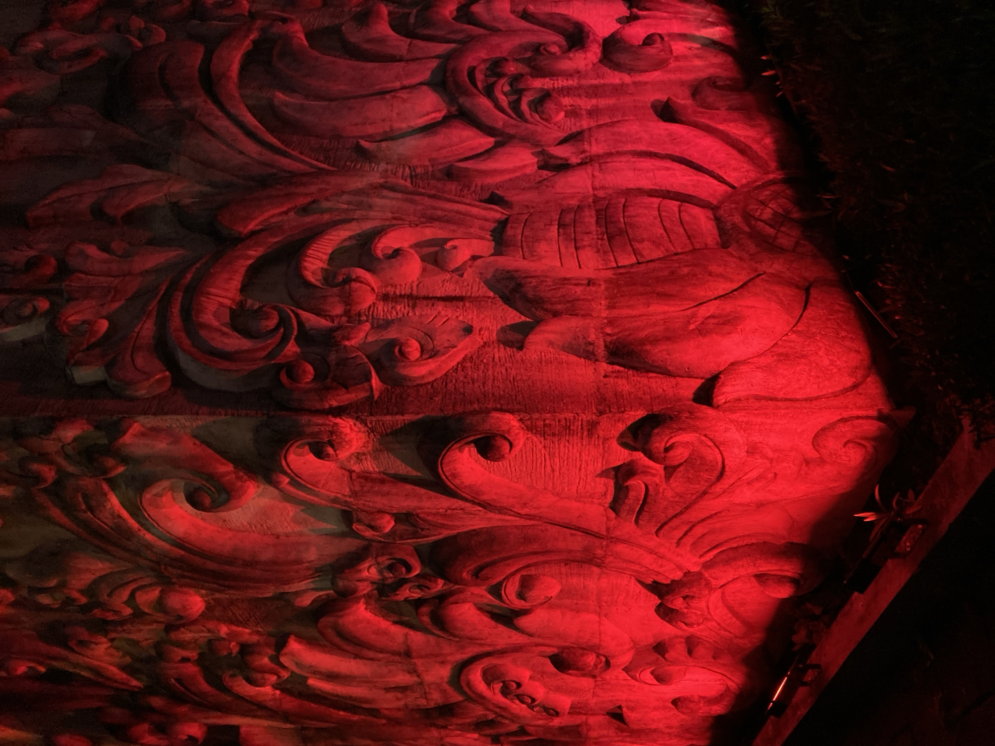 The image depicts a close-up, low-angle shot of a large, ornate stone relief illuminated by vibrant red lighting. The relief features swirling, abstract organic shapes reminiscent of foliage or waves, with a prominent vase-like structure at the center-right. The lighting casts deep shadows, emphasizing the texture and three-dimensionality of the carving. The bottom of the image shows a dark foreground with glimpses of green foliage and what appear to be ground-level lights or fixtures. The overall impression is dramatic and somewhat mysterious, with the red light creating an intense and possibly ominous atmosphere. The relief is likely a decorative element on a building or monument, given its scale and intricate design. The context of Kuta Selatan, Indonesia, suggests this could be part of a temple, resort, or public art installation, common in the region's tropical and often elaborately decorated architecture.