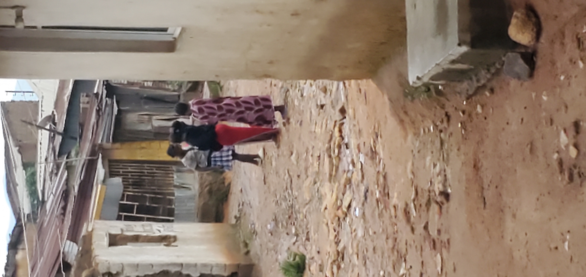 Three young people are standing in a dirt alleyway in an urban environment. One person, wearing a blue and white checkered skirt and a grey shirt, is facing away from the camera. To their right, a person in a black top and a long red skirt stands facing left. On the far right, a person in a patterned pink and purple dress stands facing left. In the background, there are several weathered buildings with corrugated metal roofs. The ground is uneven and appears to be made of packed dirt with scattered rocks and debris. The lighting suggests it is daytime, possibly overcast.