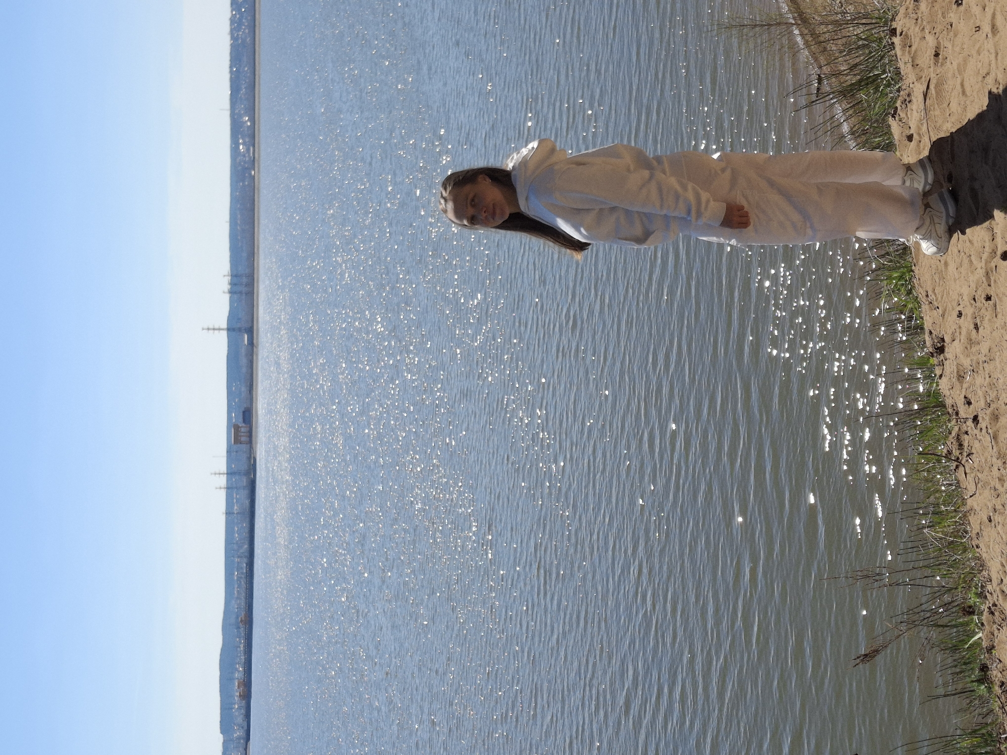 A woman wearing a white hooded sweatshirt and white pants stands on a sandy beach next to a body of water. The water is shimmering in the sunlight. In the distance across the water is the city of Noviy, Russia.