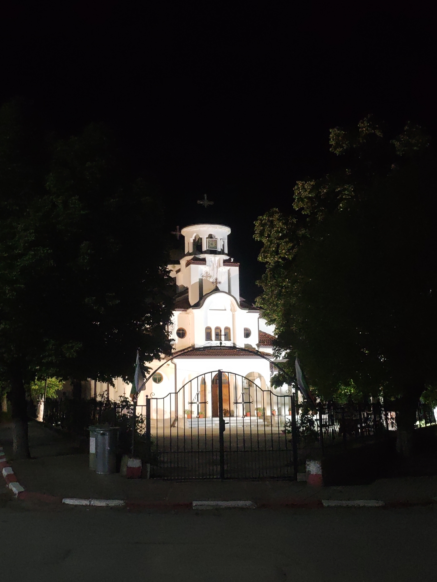 The image captures a brightly lit church at night, framed by dark trees on either side. The church features white walls, a multi-tiered tower culminating in a dome topped with an Orthodox cross, and a tiled roof. Its facade is illuminated by warm artificial lights, highlighting its architectural details, including arched windows and a decorative entrance.
In the foreground, a dark metal gate with ornate scrollwork stands closed before the church entrance. To the left of the gate, a small, light-colored bin or trash can is visible. The immediate foreground shows a paved surface, likely a road or sidewalk, with distinct red and white painted curbs or lines.
The sky is completely dark, indicating nighttime. No human activity is visible, contributing to a serene and quiet atmosphere. A second, smaller Orthodox cross is faintly visible in the upper right corner of the dark sky, possibly belonging to another structure or an extension of the church. A flag, possibly Bulgarian based on its colors, is partially visible to the left of the church entrance, behind the gate. No discernible text is visible anywhere in the image.