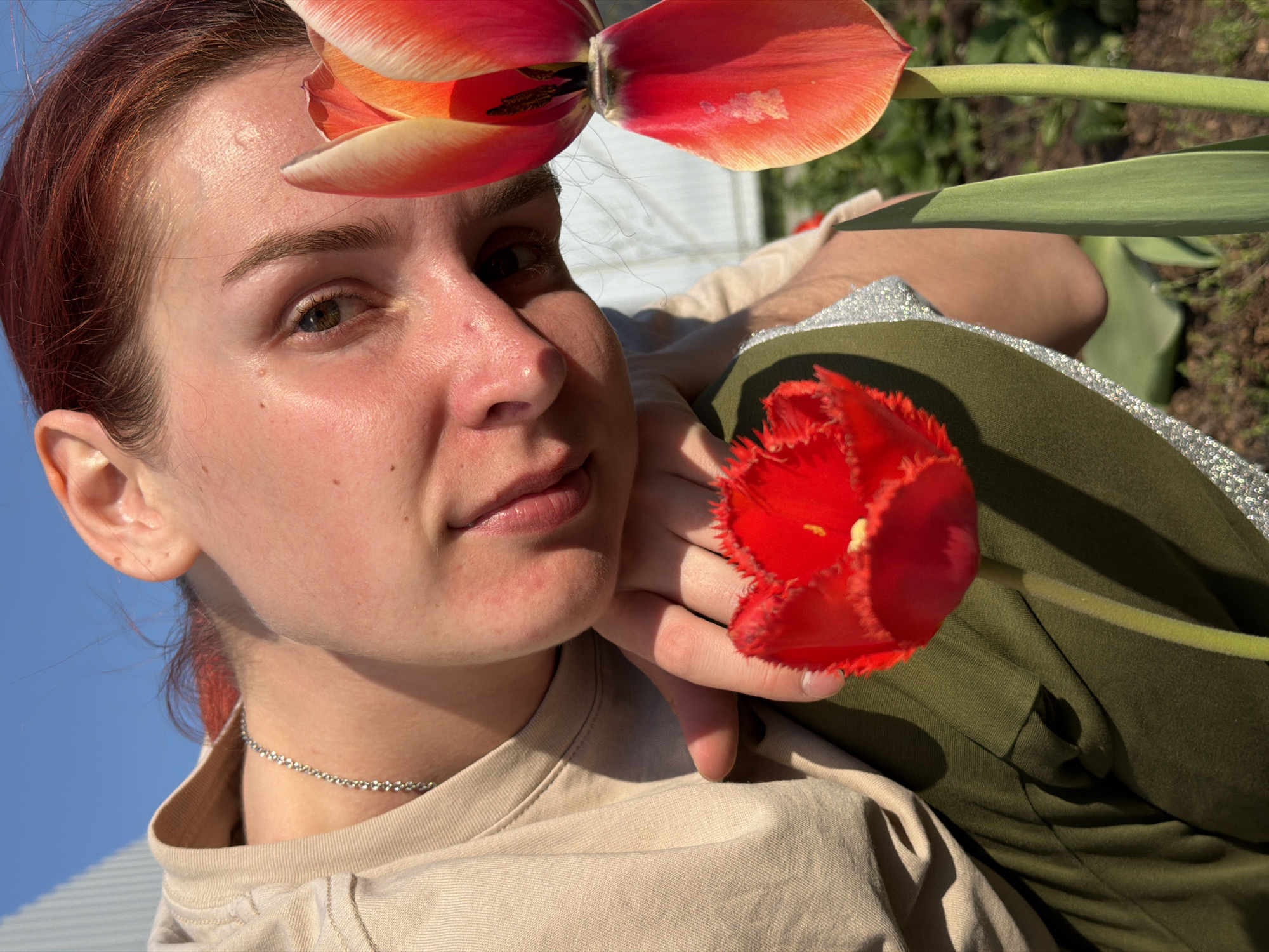 A reddish-haired person with fair skin gazes at the camera, partially obscured by two red tulips in full bloom. One tulip is positioned above their head, while the other is held close to their face. The person wears a light beige shirt and a beaded necklace. Green and shiny garments are also visible in the frame. The background features a blue sky and glimpses of a white building, with ground and foliage visible at the top right. This image appears to be taken outdoors in Chaikovsky, Russia.