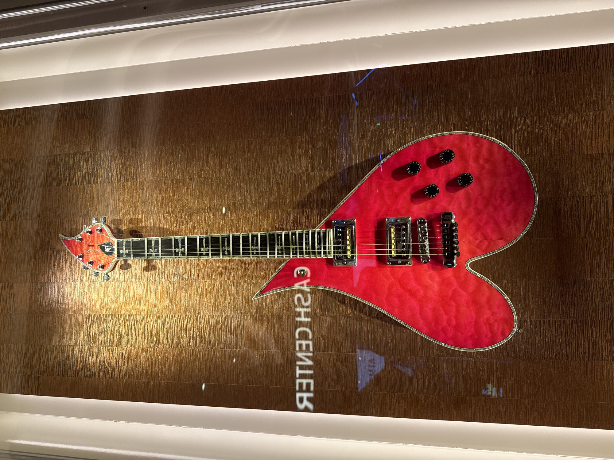 A red heart-shaped electric guitar hangs horizontally against a textured, brown-toned wall. The guitar features a headstock with tuning pegs, a fretboard with inlays, pickups, and black control knobs. The reflection on the wall contains the text "CASH CENTER" reversed. The scene is illuminated by both direct and reflected light. It is located in the city of Hollywood Seminole Reservation, United States of America.