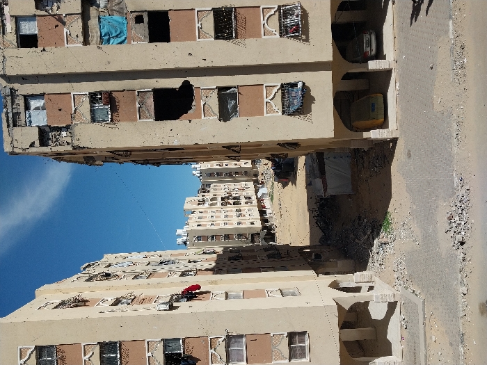 The image depicts a residential urban environment in Khan Yunis, Palestinian Territory, under a clear blue sky with minimal cloud cover. Two multi-story, light beige apartment buildings, exhibiting varying degrees of damage, frame the central view.
The building on the left presents a relatively intact facade with some sections showing deterioration and missing plaster. Several windows are visible, some open, some with glass, and a red piece of clothing hangs from a lower-level window. Its ground floor features arched open bays.
The building on the right shows more extensive damage, including a large, irregular, dark cavity on a central floor, indicative of an impact. Numerous smaller impact marks (pockmarks) are visible across its exterior. Multiple windows are broken, missing glass, or covered with fabric, notably a blue sheet on a middle floor. The ground floor also incorporates arched open bays, where a yellow covered vehicle and a white sedan are parked.
The ground in the immediate foreground is a partially paved surface with broken pavers and scattered debris. Between the two main buildings, the ground transitions to an unpaved, sandy, and rubble-filled area. Twisted metal, construction debris, and discarded items are scattered throughout this passage. A small, temporary structure, possibly a tent or shelter, with a light-colored tarp roof, is situated within this central passage. Green vegetation patches emerge from the debris. In the background, further multi-story apartment buildings, similar in style, are visible, separated from the foreground structures by a barren, unpaved expanse. Thin wires are stretched horizontally across the sky between the foreground buildings. No individuals are present in the scene.