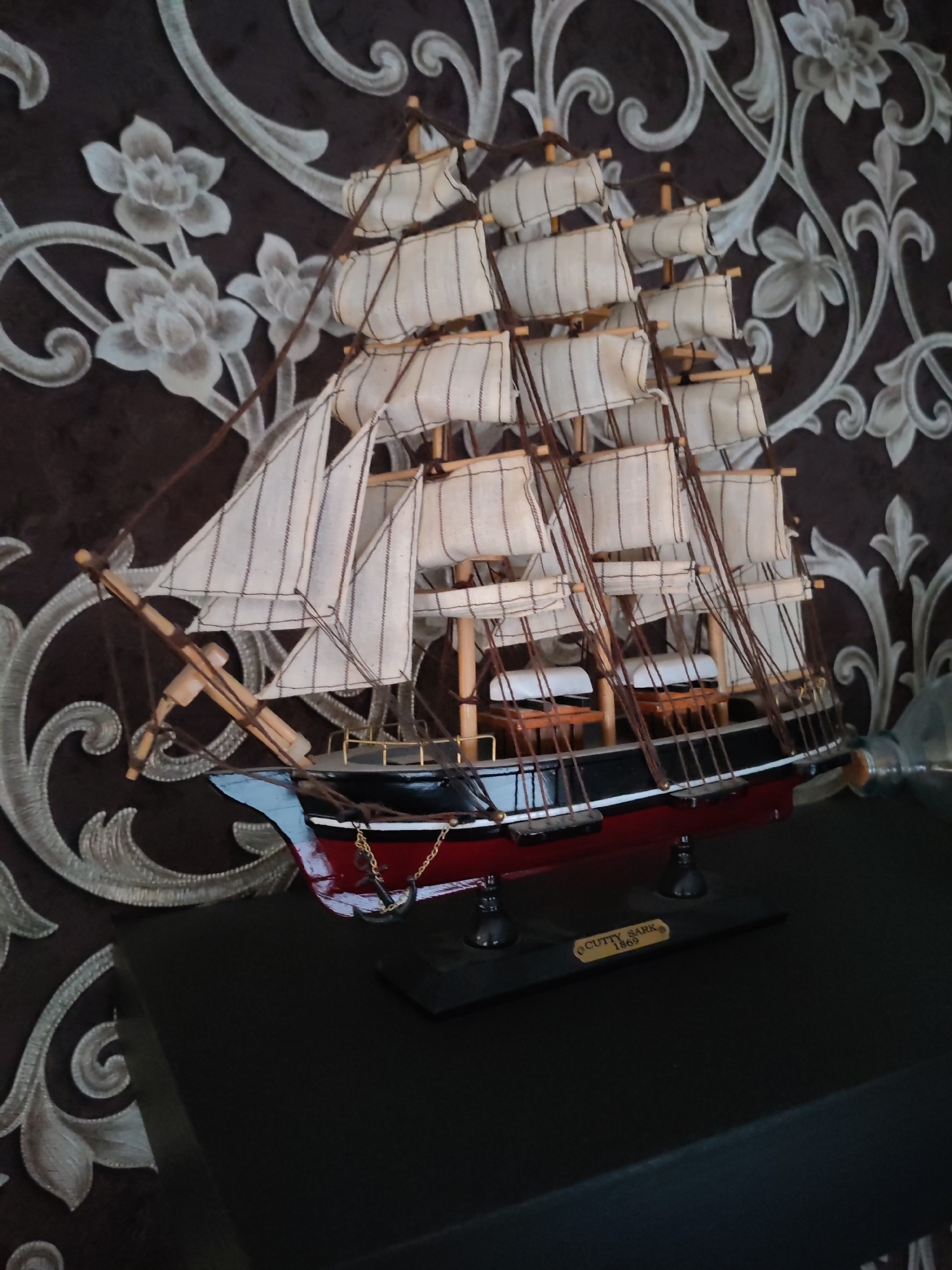 A model ship, identified as the "Cutty Sark 1869," is displayed on a dark, flat surface in front of a dark brown damask wallpaper with a floral pattern. The ship is predominantly red and black with white sails. The model is detailed, featuring masts, rigging, and small benches on its deck. The wallpaper exhibits a repeating pattern of stylized flowers and swirling decorative elements in a lighter, possibly silver or gray, color. The overall lighting suggests an indoor setting, possibly a home environment. No individuals or actions are depicted. The image does not provide any information about the city of Bol'shie Mkhi, Russia.