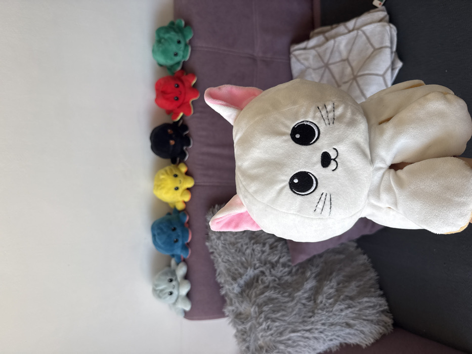 An indoor scene in Plovdiv, Bulgaria, depicts a collection of plush toys on and around a sofa against a white wall. In the immediate foreground, a white plush cat, featuring embroidered black eyes, a black nose, and whiskers, with light pink inner ears, is centrally positioned and partially held from below. Behind the cat, a purple fabric sofa is visible. On the left side of the sofa, a large grey, shaggy throw pillow rests. On the right side, a white blanket with a grey geometric pattern is folded. Along the backrest of the sofa, from left to right, six smaller reversible octopus plush toys are arranged in a row: light grey, blue, yellow, black, red, and green. The floor or a low surface beneath the sofa appears dark.