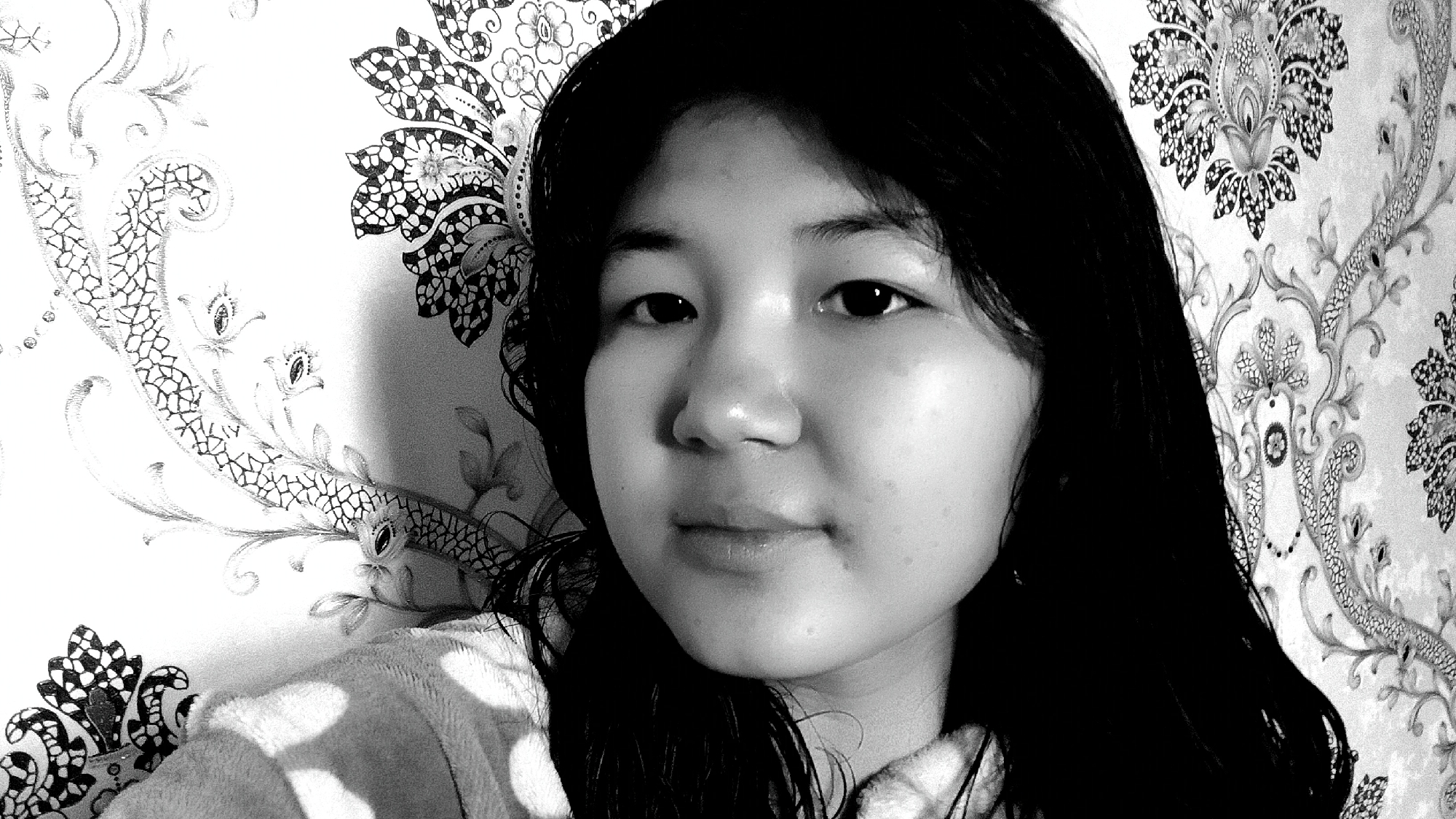 In a black and white medium shot, a young woman with dark, straight hair is positioned slightly off-center to the right, looking directly at the camera. Her face is illuminated from the left, casting a shadow on the right side of her face and hair. She is wearing a light-colored, fluffy garment, likely a robe or sweater, visible on her left shoulder and chest.
The background is a patterned wallpaper featuring an intricate, ornate design with floral and paisley-like motifs. The pattern is in shades of grey and black against a lighter background. To the left of the woman, a segment of the wallpaper shows a stylized, serpentine design with scale-like textures and what appear to be small, eye-like elements.
There are no discernible elements in the image that place the scene in the city of Uchkun, Kyrgyzstan. The image appears to be a self-portrait taken indoors, with the focus being on the woman and her immediate surroundings.