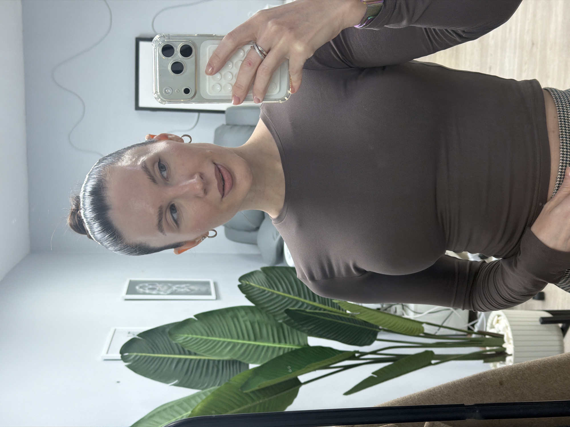 A fair-skinned woman with brown hair pulled back in a bun takes a selfie with a white-encased iPhone. She wears a long-sleeved brown shirt and a watch on her left wrist. Her left hand holds the phone with a ring on her ring finger. The background includes a gray wall, two framed pictures, a green plant, a gray couch, and a window with a white cord hanging down. The location is presumably Perm, Russia, based on visual analysis.