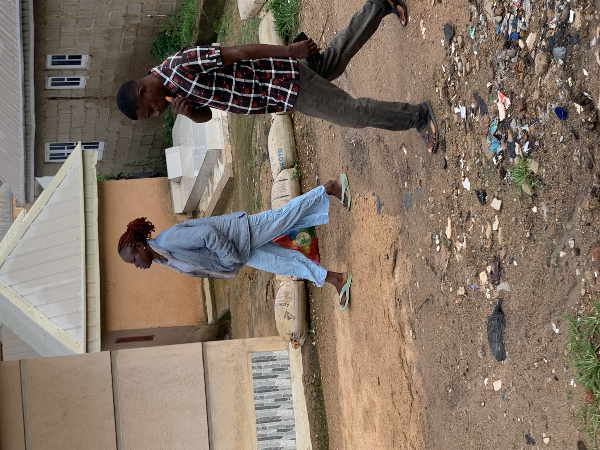 The image captures a street scene in Jos, Nigeria, with two individuals walking on a dirt path. A woman with red braided hair, wearing a light blue puffer jacket and light blue pants, is seen walking from left to right. She is wearing green flip-flops and carrying a colorful bag in her left hand. To her right and slightly behind her, a man with short dark hair, dressed in a red and black checkered short-sleeved shirt and dark pants, walks in the same direction. He is holding a small orange object to his mouth and a dark rectangular object in his right hand. The ground is a mix of dirt and scattered debris, including plastic fragments and paper. In the background, there are buildings constructed with concrete blocks and zinc roofs, typical of residential areas in Jos. Several large sacks, possibly containing cement, are also visible on the ground. The overall impression is one of an everyday urban environment with signs of human activity and the presence of litter.