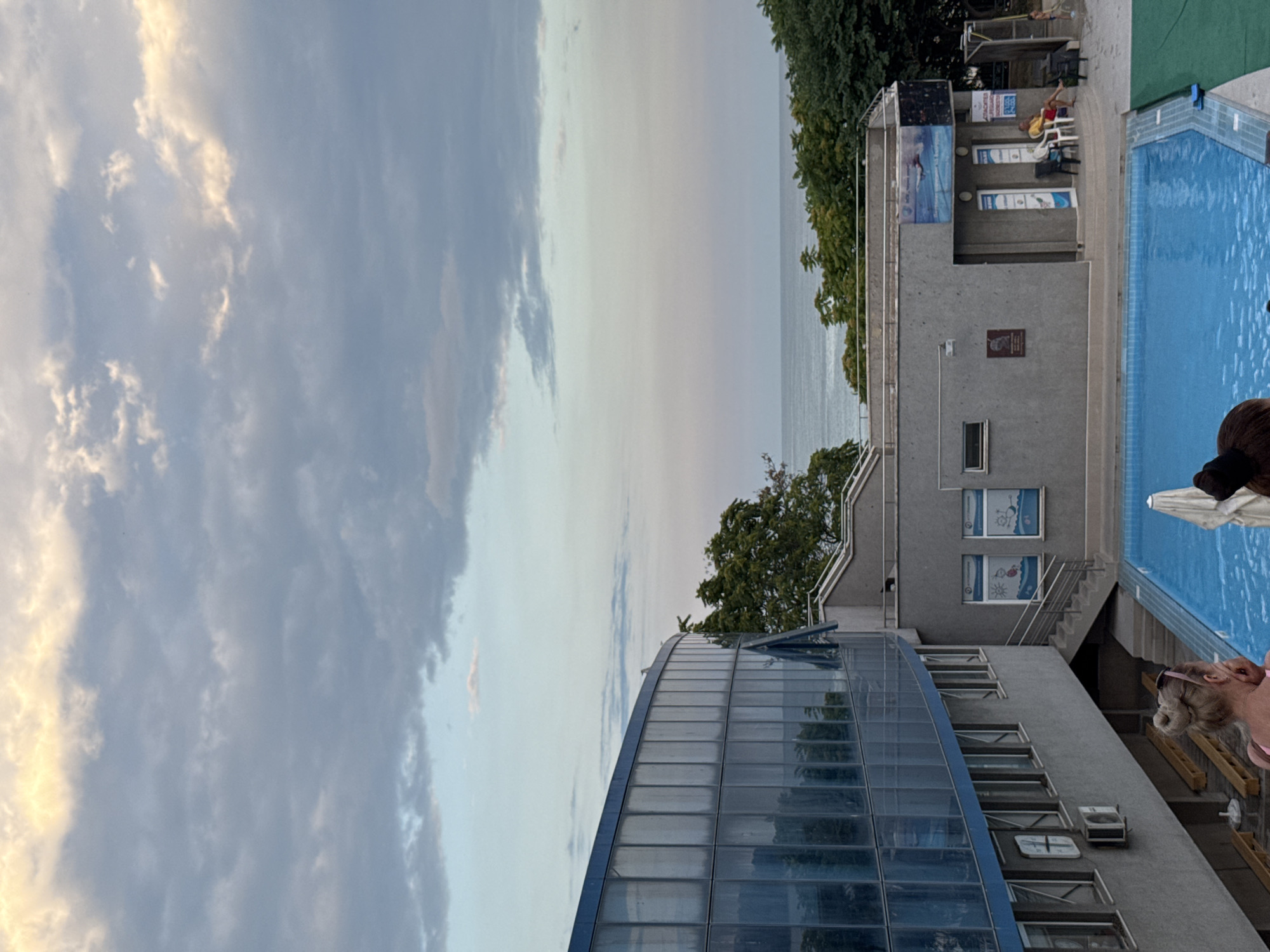 The image captures a poolside scene at a resort or public facility in Varna, Bulgaria. In the foreground, the vibrant blue water of a swimming pool occupies the lower portion of the frame. On the left, a modern building with a curved facade clad in blue glass windows stands prominently. Adjacent to it, a gray, multi-story building features a staircase leading to an elevated area. Several people are visible: a woman with blonde hair in a bun, wearing a pink top, is partially seen in the lower left corner, looking towards the pool. Further back, on the elevated section of the gray building, a person in a yellow shirt sits on a chair, and other individuals are scattered around, including a child near a shower area. The background reveals a dense line of green trees, beyond which lies the calm, gray expanse of the sea under a sky filled with dramatic clouds, illuminated by warm, soft light, suggesting either dawn or dusk. The overall atmosphere is tranquil, with the resort facilities and the natural elements of the sea and sky harmonizing.